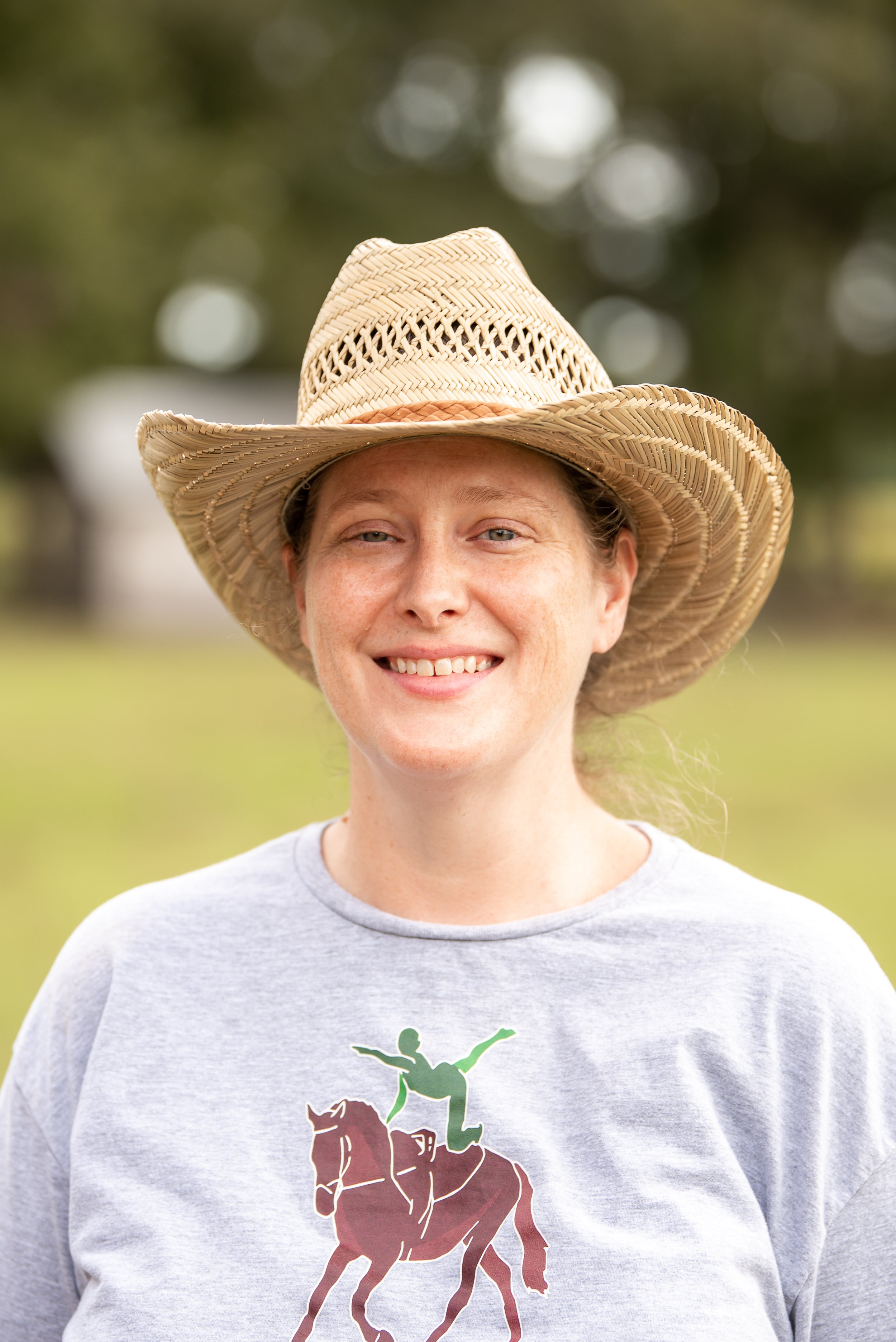 A woman smiling outdoors wearing a straw hat and a gray T-shirt with a graphic of a person riding a horse.