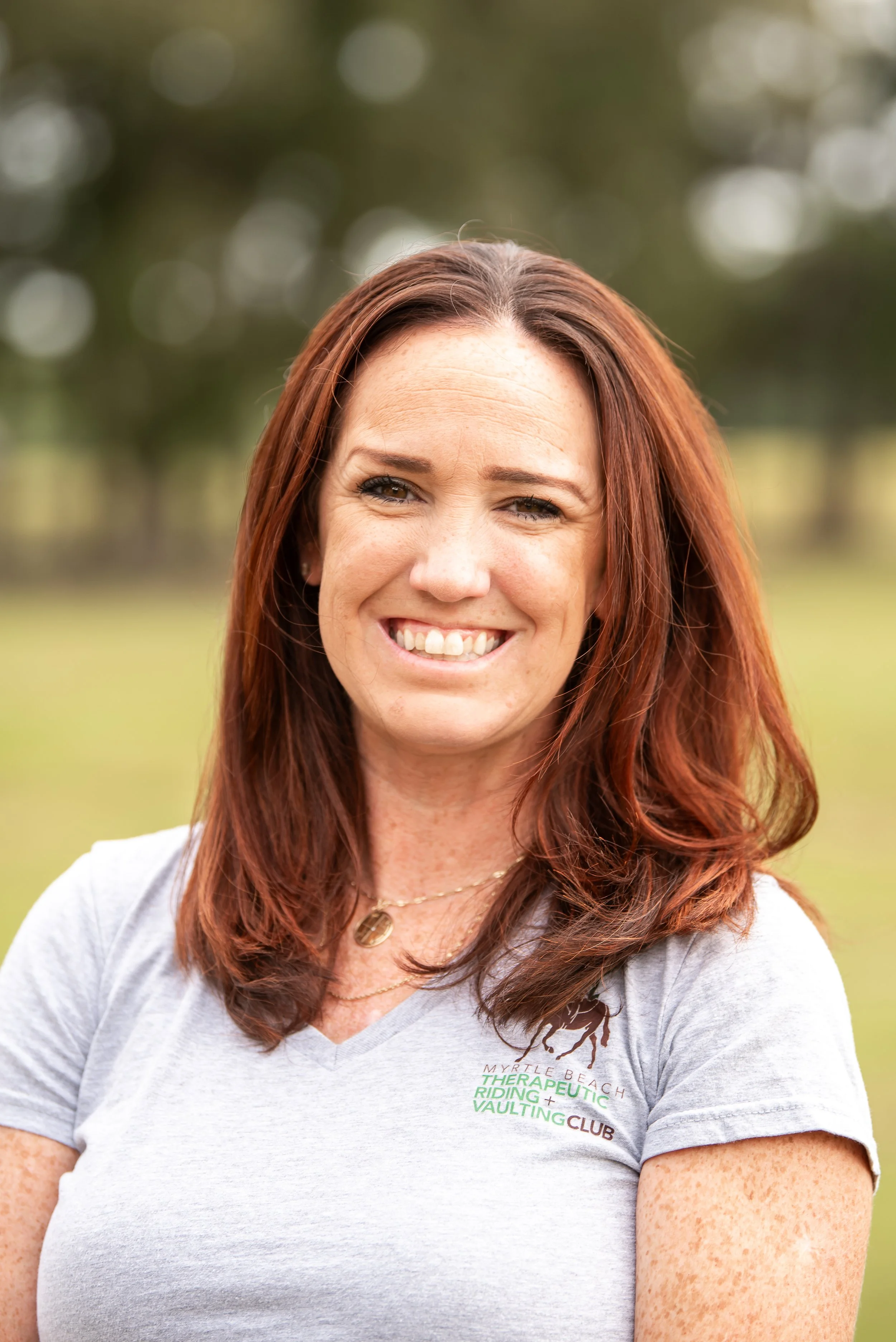 A woman with shoulder-length red hair smiling outdoors, wearing a light gray T-shirt with a logo and gold necklace.