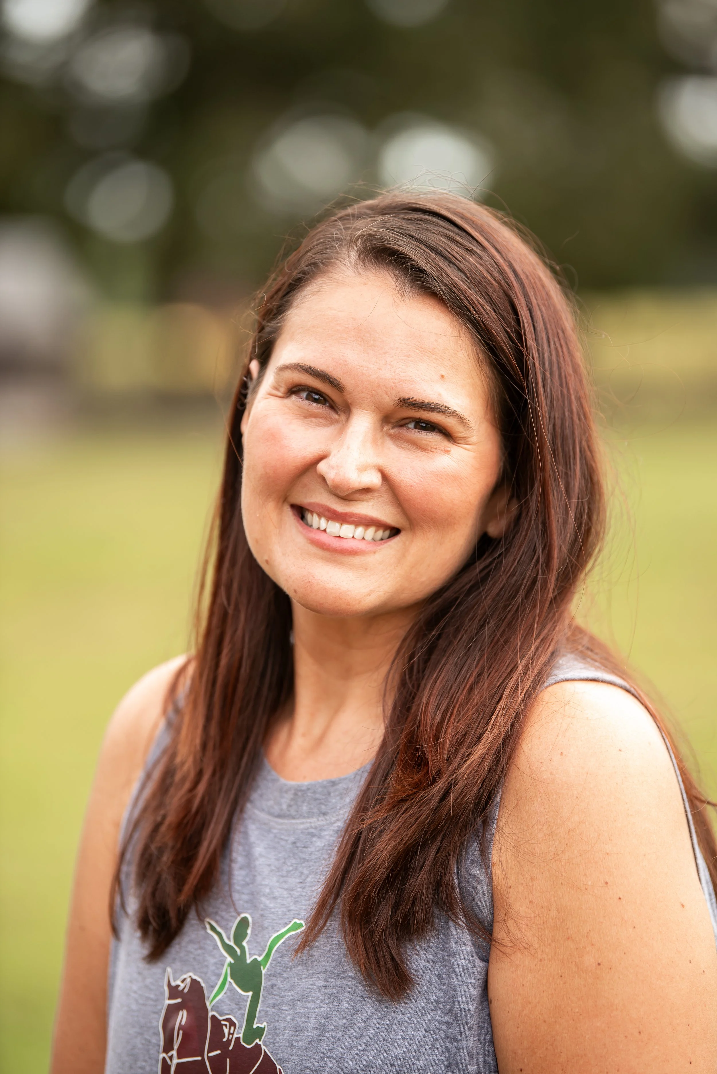 Smiling woman with long brown hair outdoors in a park, wearing a gray sleeveless top.