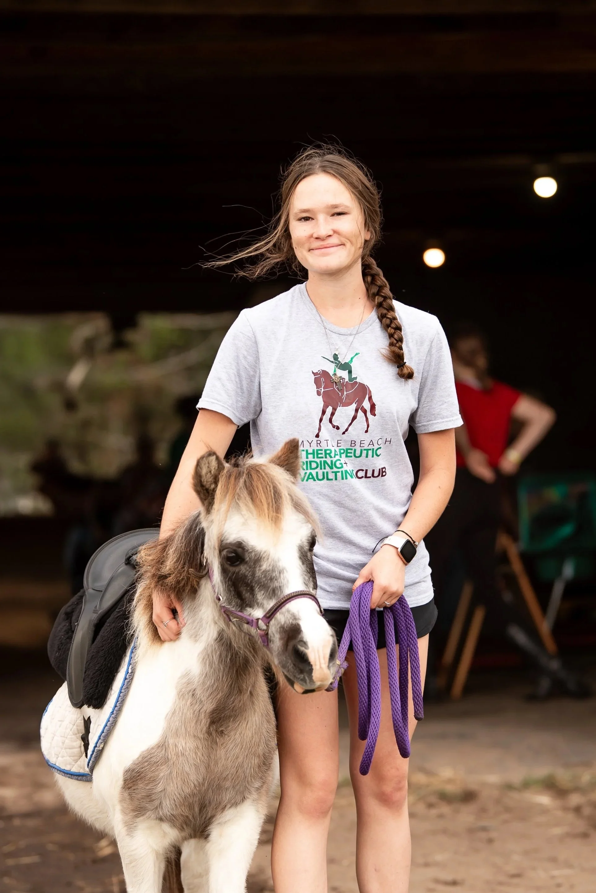 A young woman with a braid smiling and holding a purple lead rope, standing next to a small horse or pony with a saddle and bridle, in an indoor riding arena.