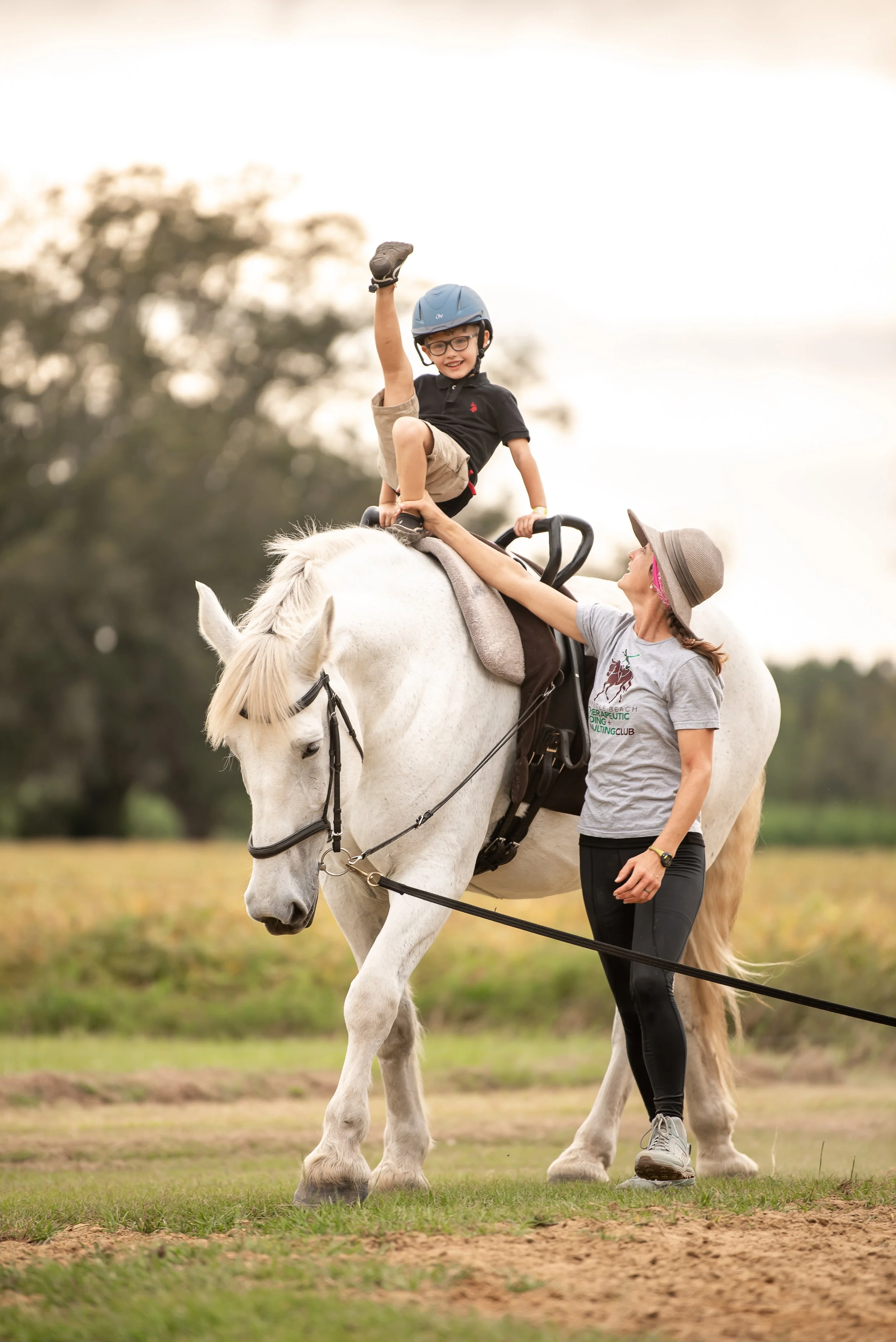 A young boy in a black shirt, beige shorts, and a helmet riding a white horse, raising his hand in the air, with a woman in a gray shirt and wide-brimmed hat holding onto the boy's leg, outdoors on a grassy field.