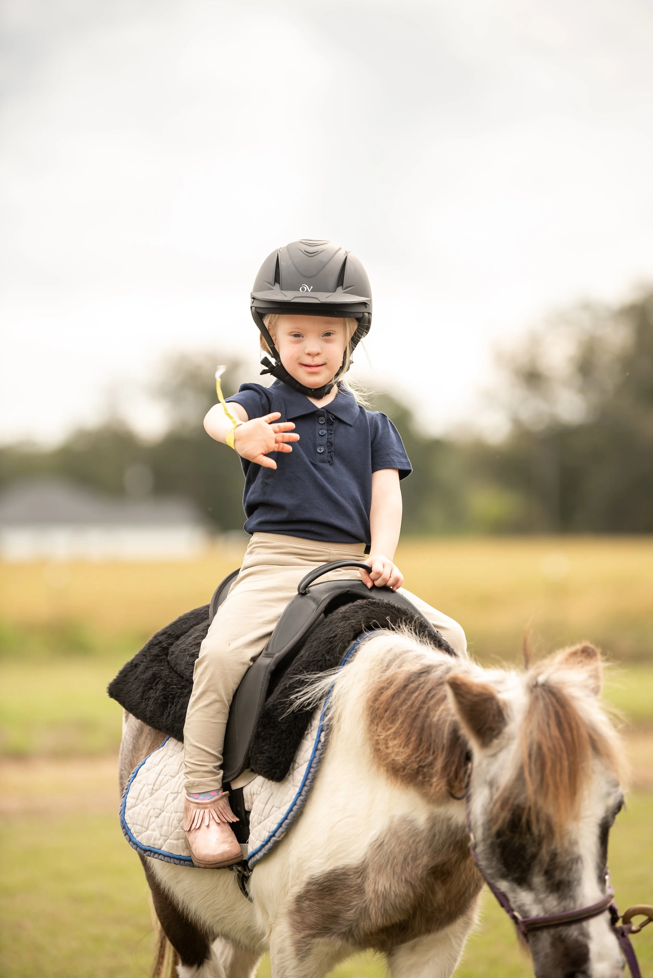 A young girl riding a pony on a grassy field, wearing a black helmet and navy polo shirt, waving at the camera.