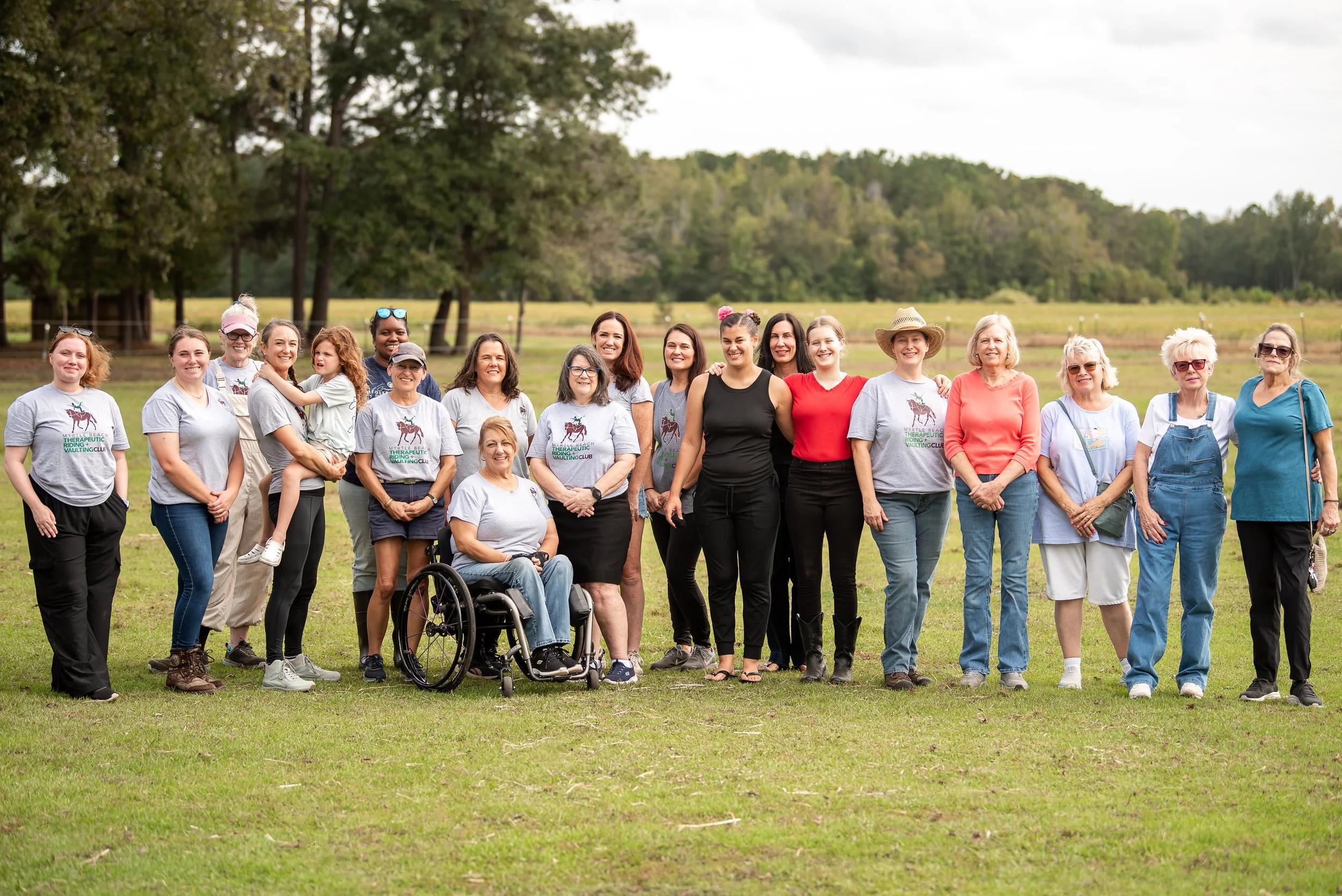 Group of women standing outdoors on green grass with a lake and trees in the background. Some women are wearing gray T-shirts with a horse and text, others are in casual clothing. There is one woman in a wheelchair, and a woman is holding a young girl.