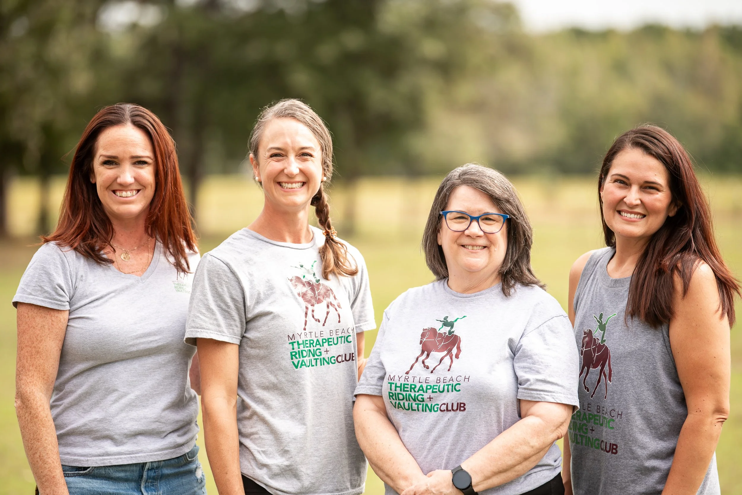 Four women standing outdoors on grass, smiling, wearing matching gray T-shirts with a horse and rider graphic, with a background of green trees and sky.