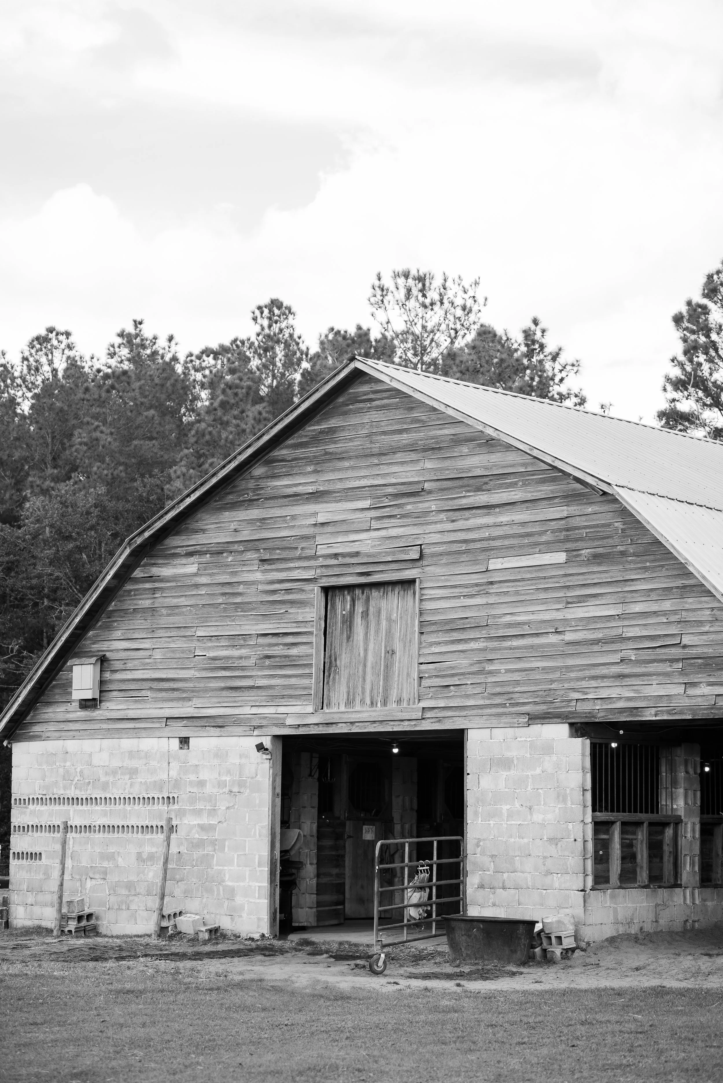 Black-and-white photo of a rustic barn with a wooden upper section and a brick lower section, open entrance, and surrounded by trees and grass.