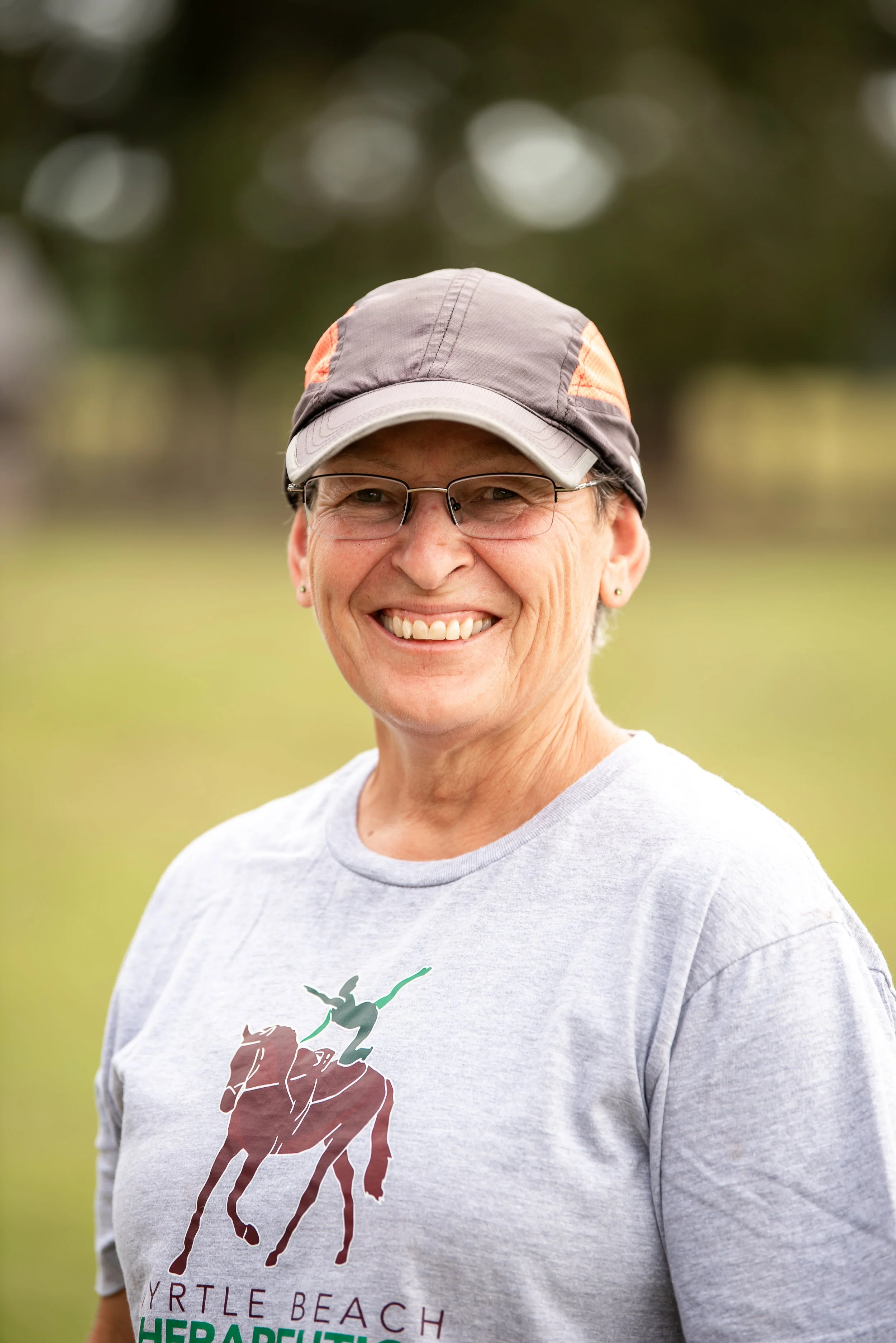 Smiling woman wearing glasses, a gray baseball cap, and a gray T-shirt with a logo of a horse racing and a person on a horse, standing outdoors with a blurred green background.