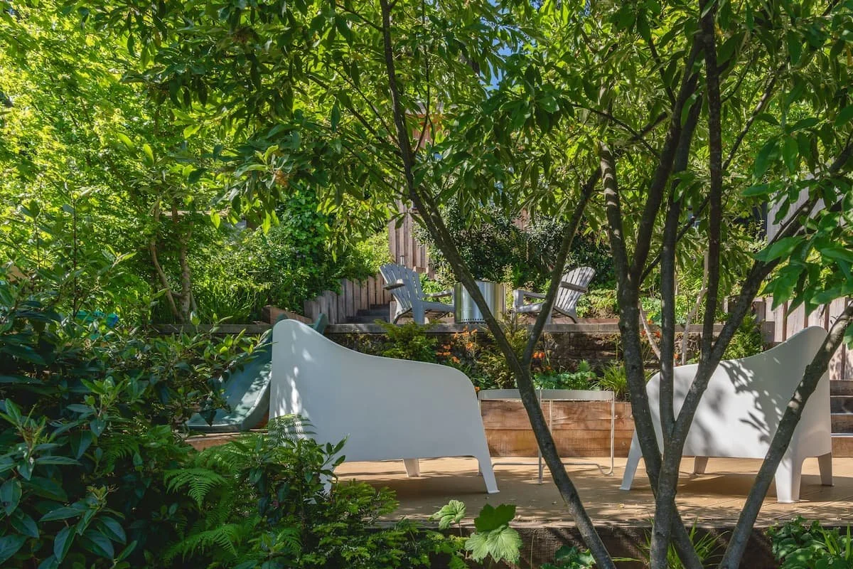 A cozy outdoor garden seating area with modern white benches, surrounded by lush green plants and trees, with a raised wooden deck in the background featuring Adirondack chairs and a small table.