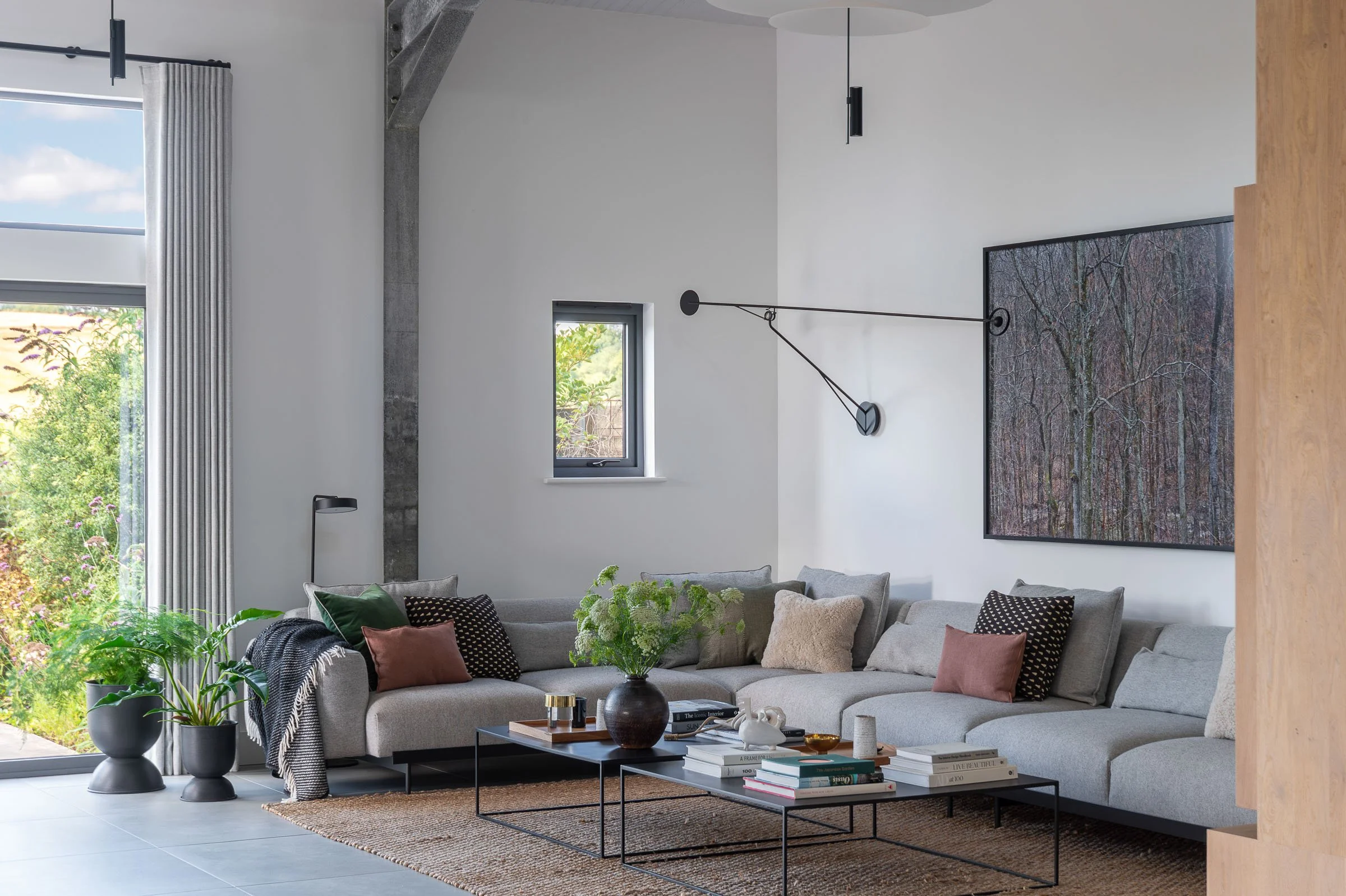 Modern living room with a large gray sectional sofa, assorted pillows, a black coffee table with books and a vase, a plant, a small window, large glass door, curtain, gallery wall art, and minimalistic lighting fixtures.