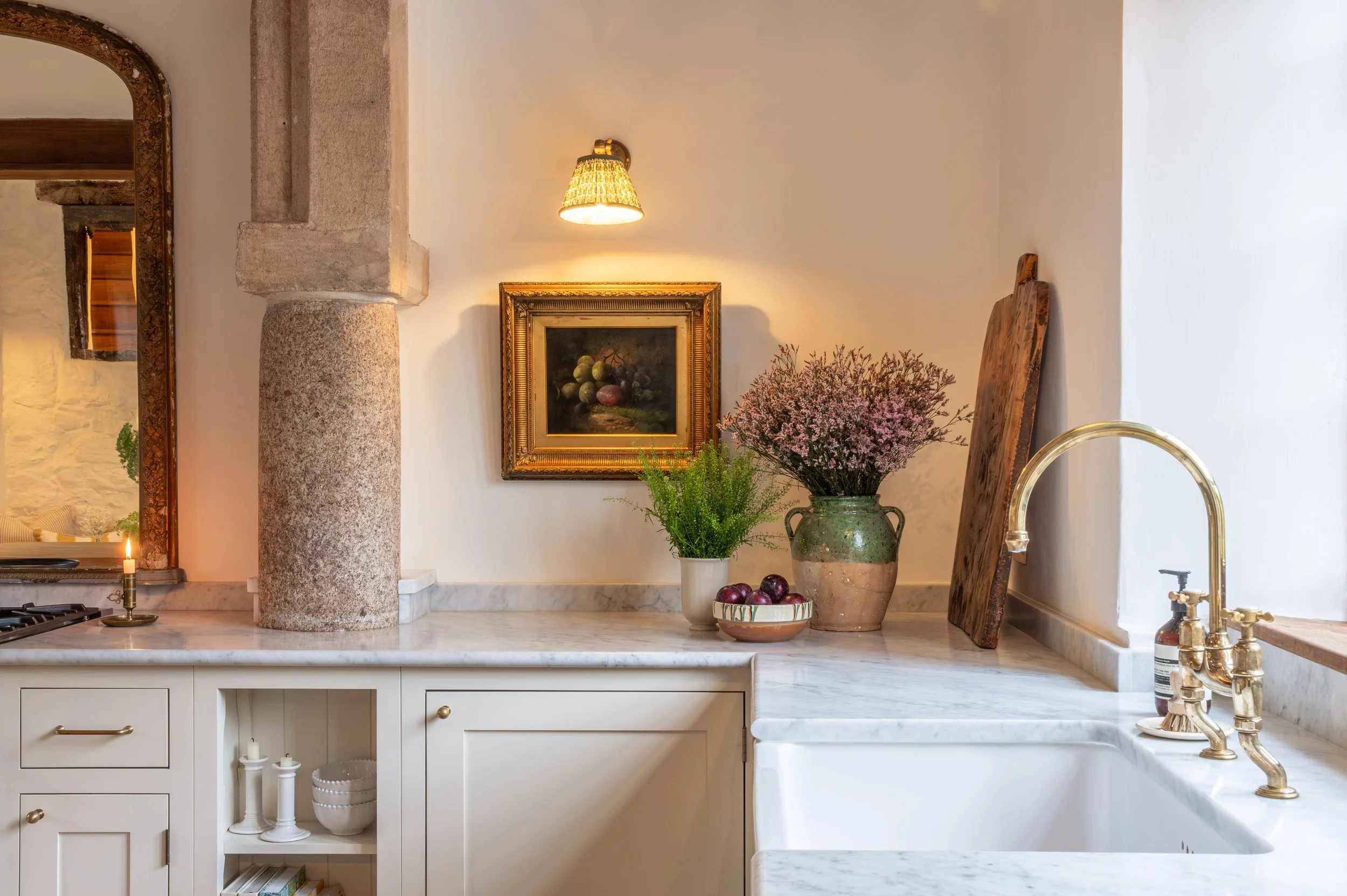 A kitchen with a white marble countertop, brass faucet, and various decorative items including a large vase with pink flowers, another with green foliage, a bowl of fruit, a wooden cutting board, and a framed still life painting. There is a wall-mounted light above the painting.