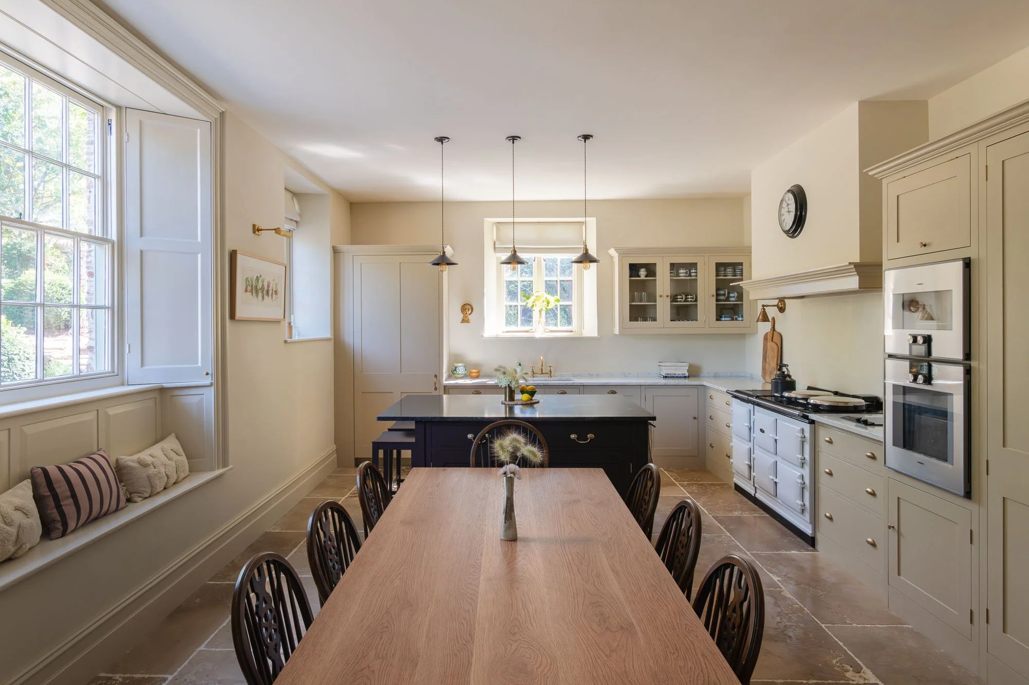 A kitchen countertop with a marble surface, a gold faucet, a large green flower vase with pink flowers, a smaller green plant, a bowl with dark red apples, a rustic wooden cutting board, a small framed painting of fruit, and a wall-mounted light fixture over the painting.