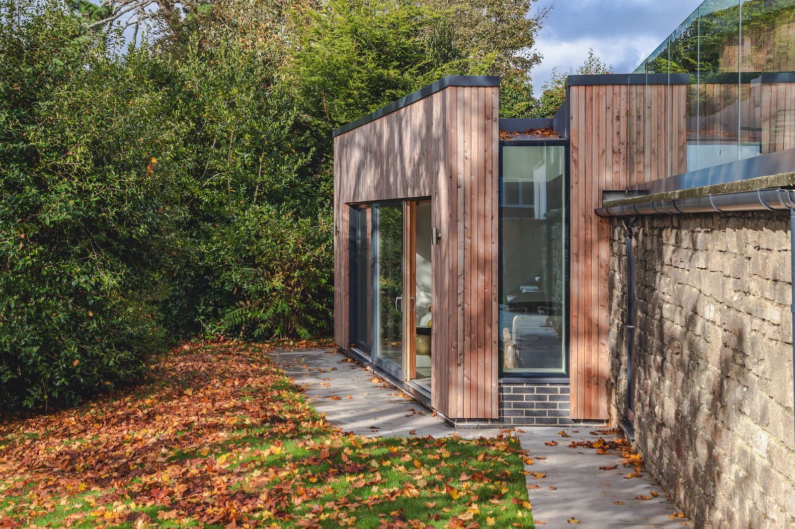 Modern small building with wooden and glass exterior, located beside an old stone wall. The building features large glass windows and a sliding door, with autumn leaves scattered on the grassy area and pathway in front.