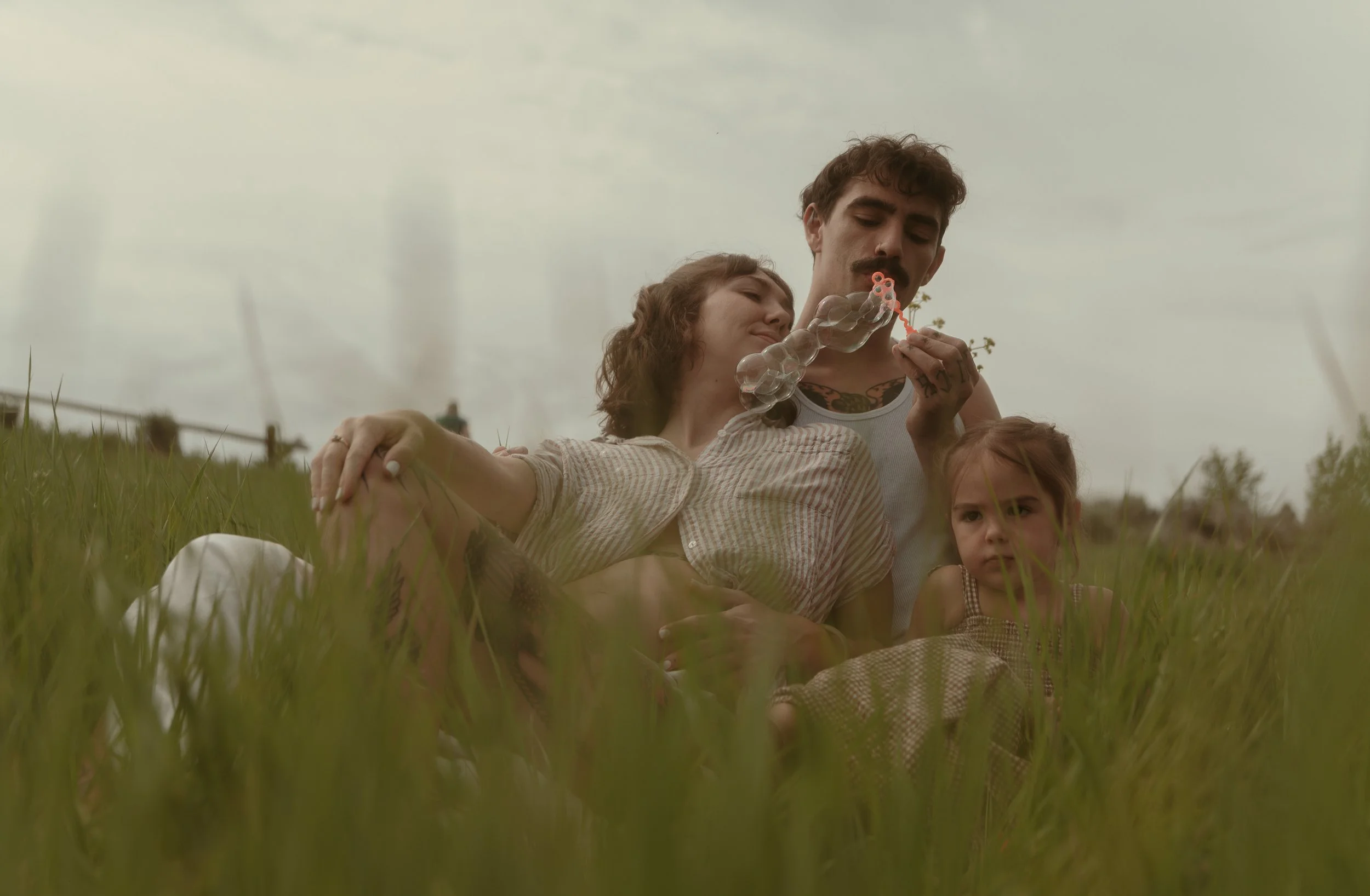 A family of three sitting in a grassy field on a cloudy day, with a man blowing bubbles, a woman leaning on him, and a girl sitting nearby watching.
