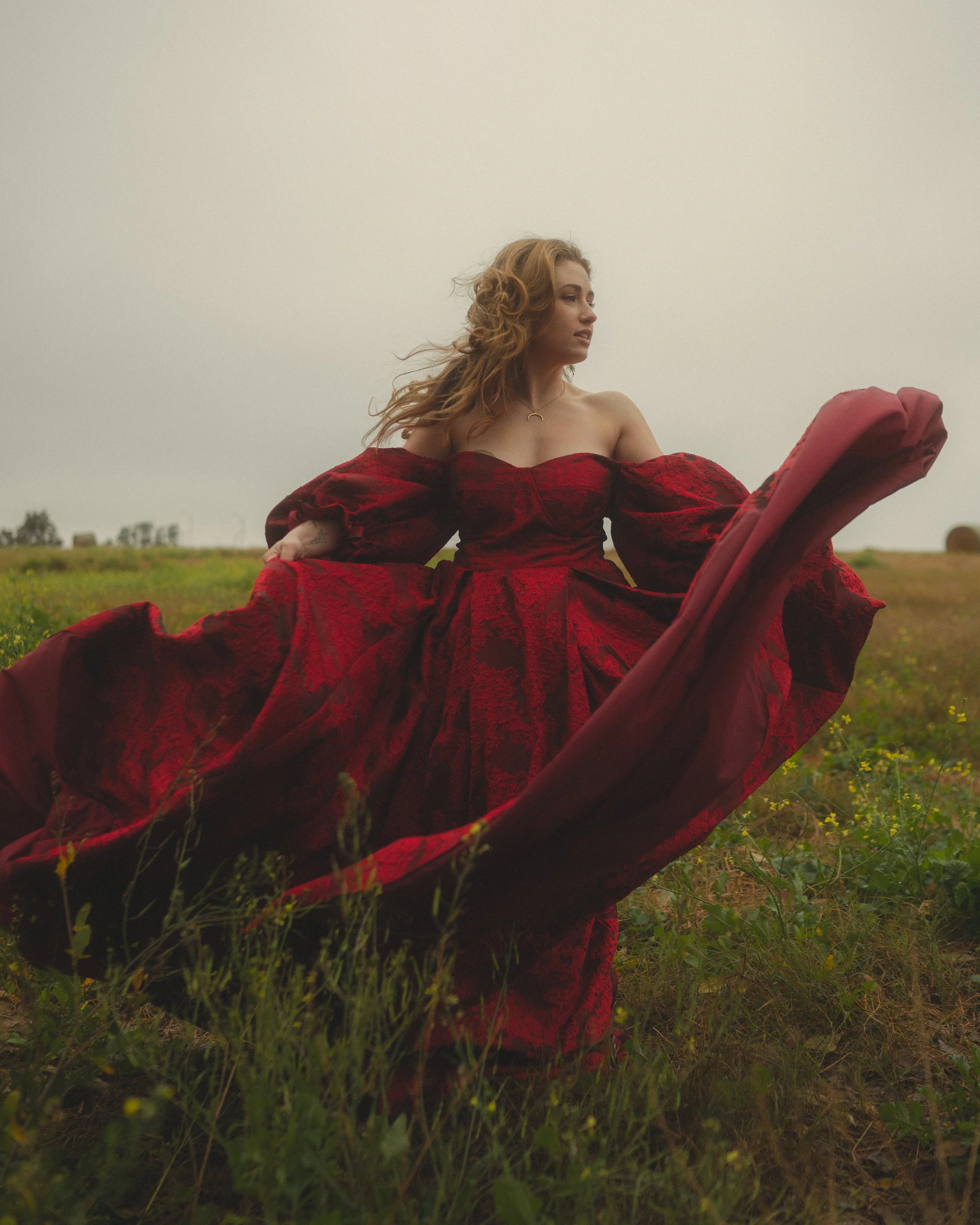 Woman in a red dress standing in a field with tall grass and yellow flowers, overcast sky, looking to the side.