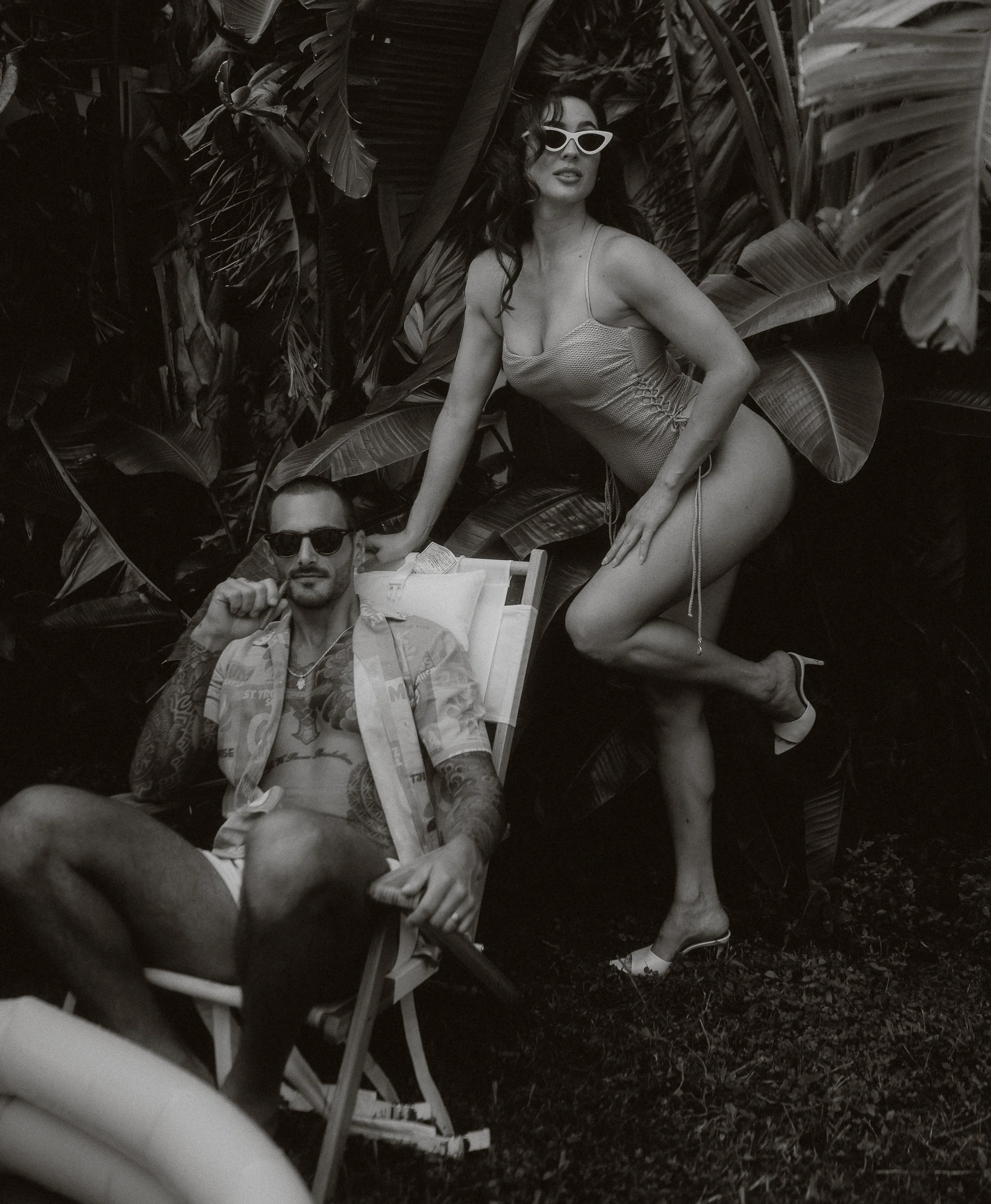 A woman in a one-piece swimsuit and sunglasses kneels next to a man with tattoos, sunglasses, and an open shirt, sitting in a deck chair among tropical plants.