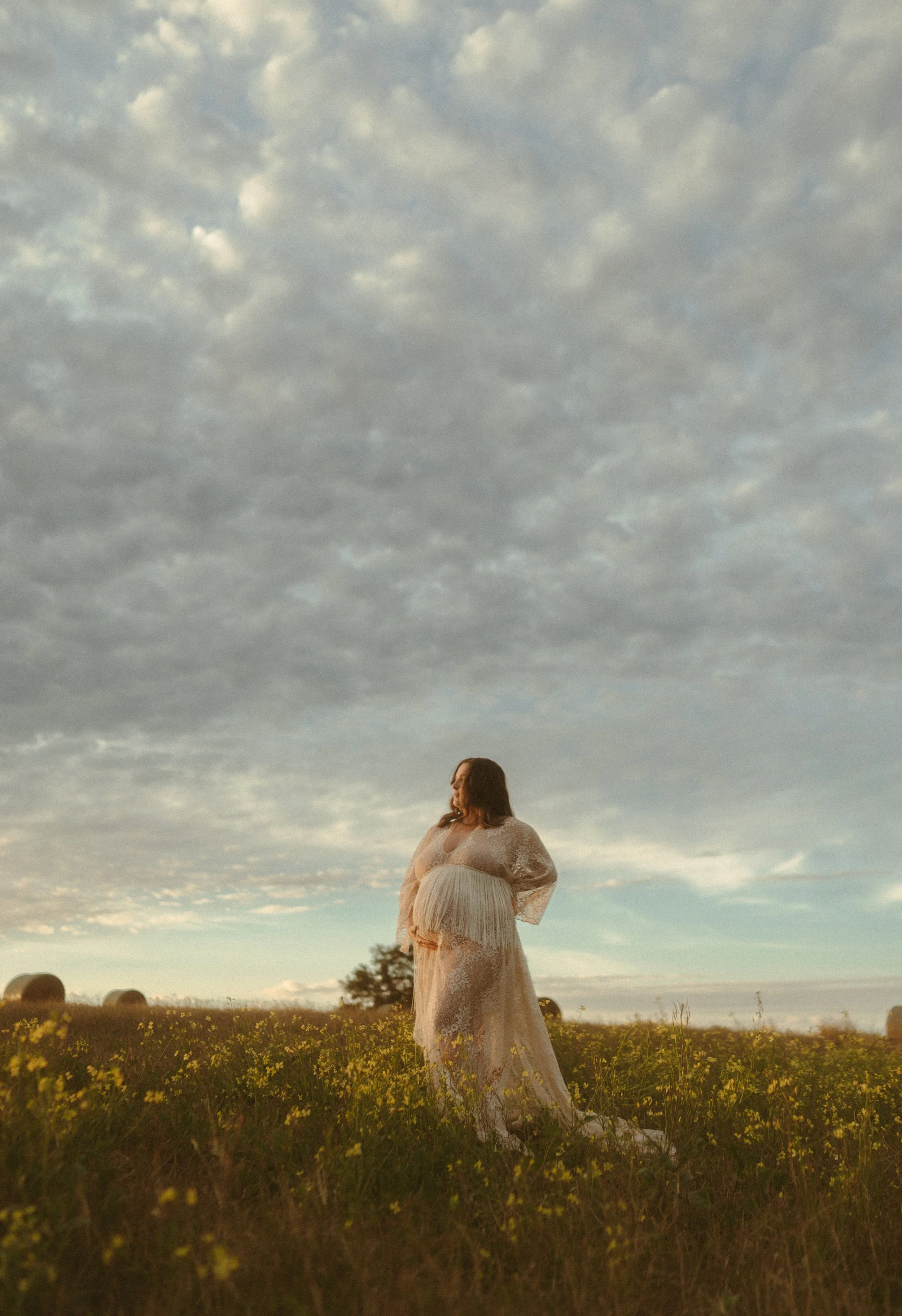 A pregnant woman standing in a field of yellow flowers at sunset, wearing a lace dress.