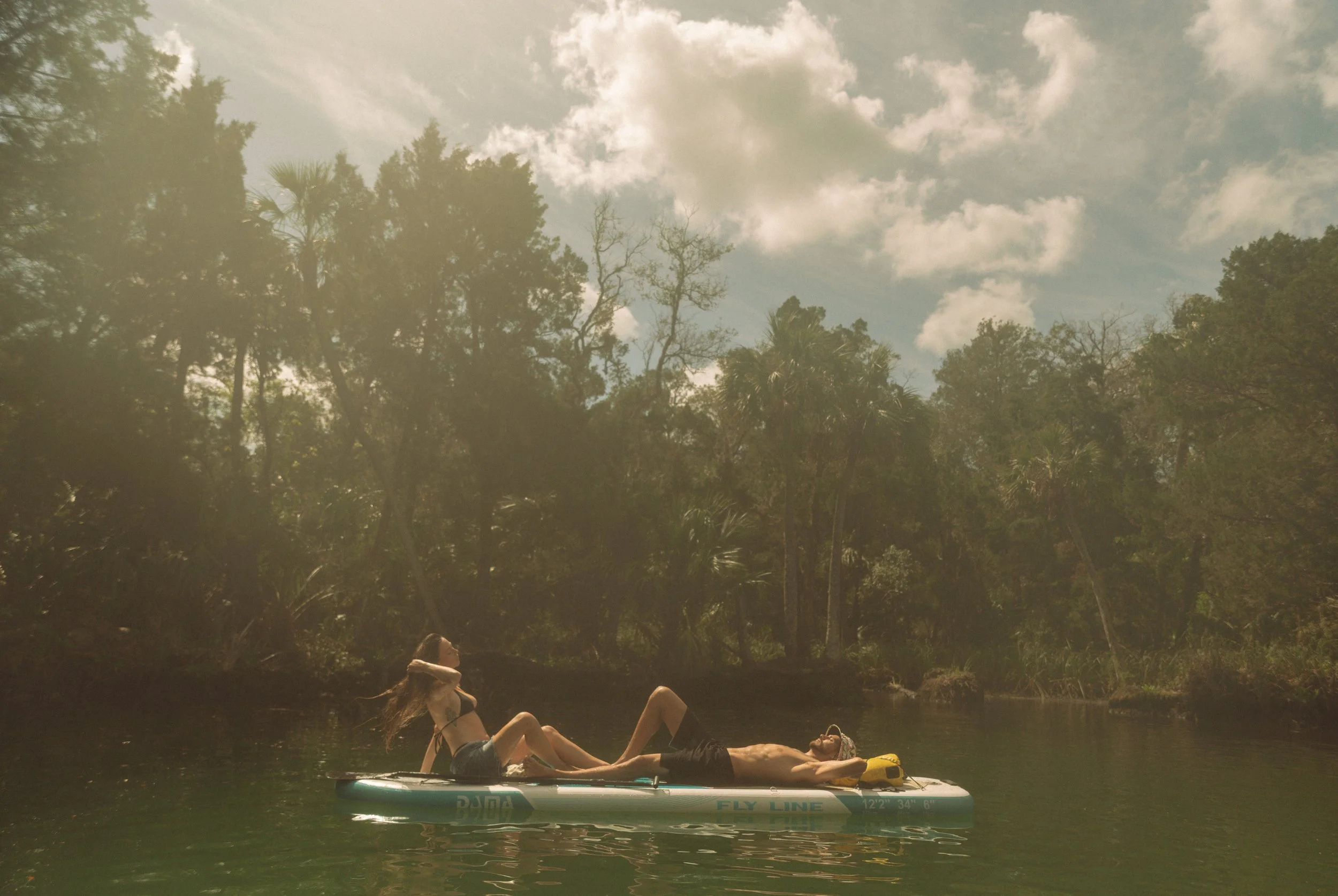 Two people relaxing on a paddleboard on a calm river surrounded by lush trees under a partly cloudy sky.