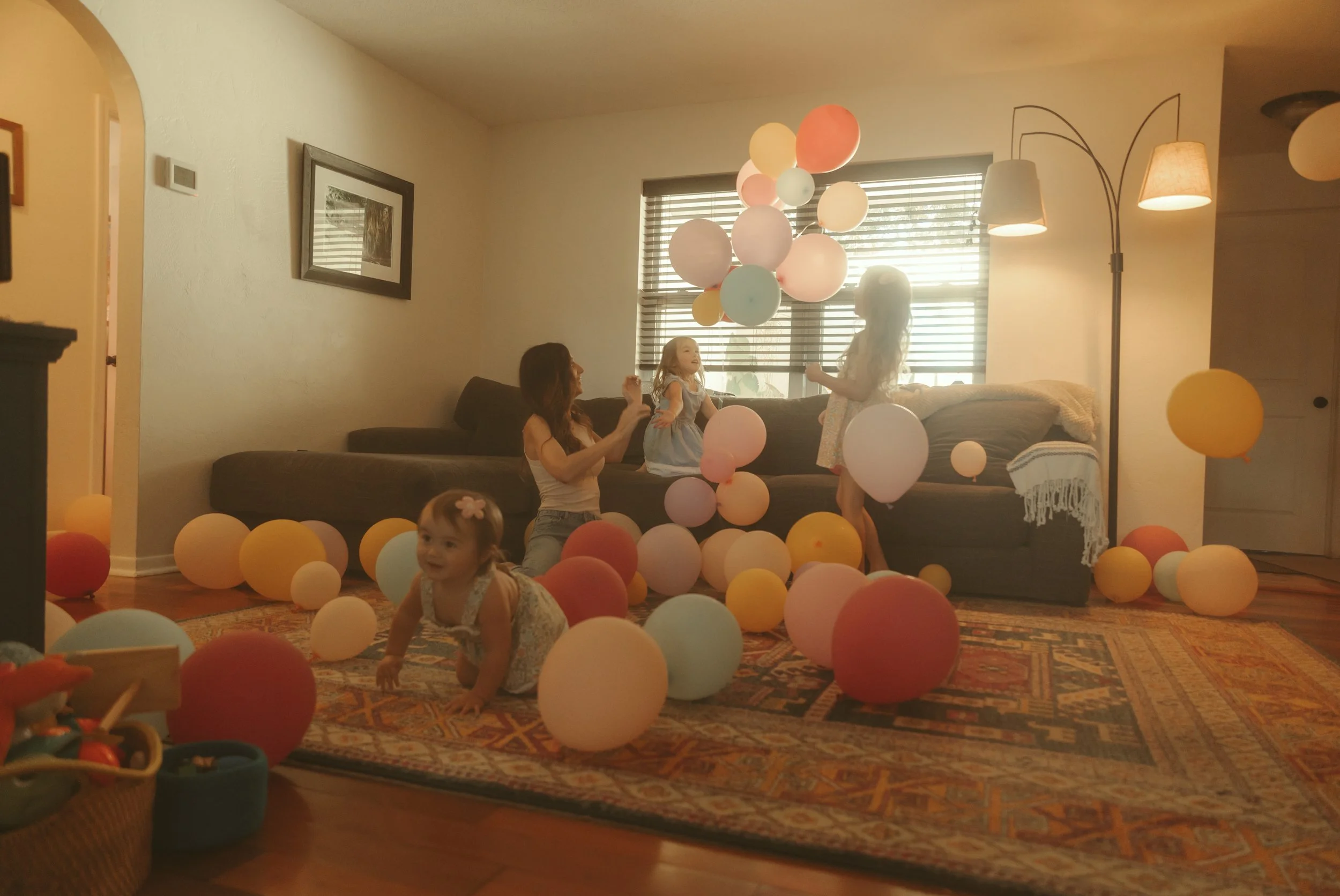 Children playing with colorful balloons in a living room with warm lighting, a large window with blinds, and a patterned rug.