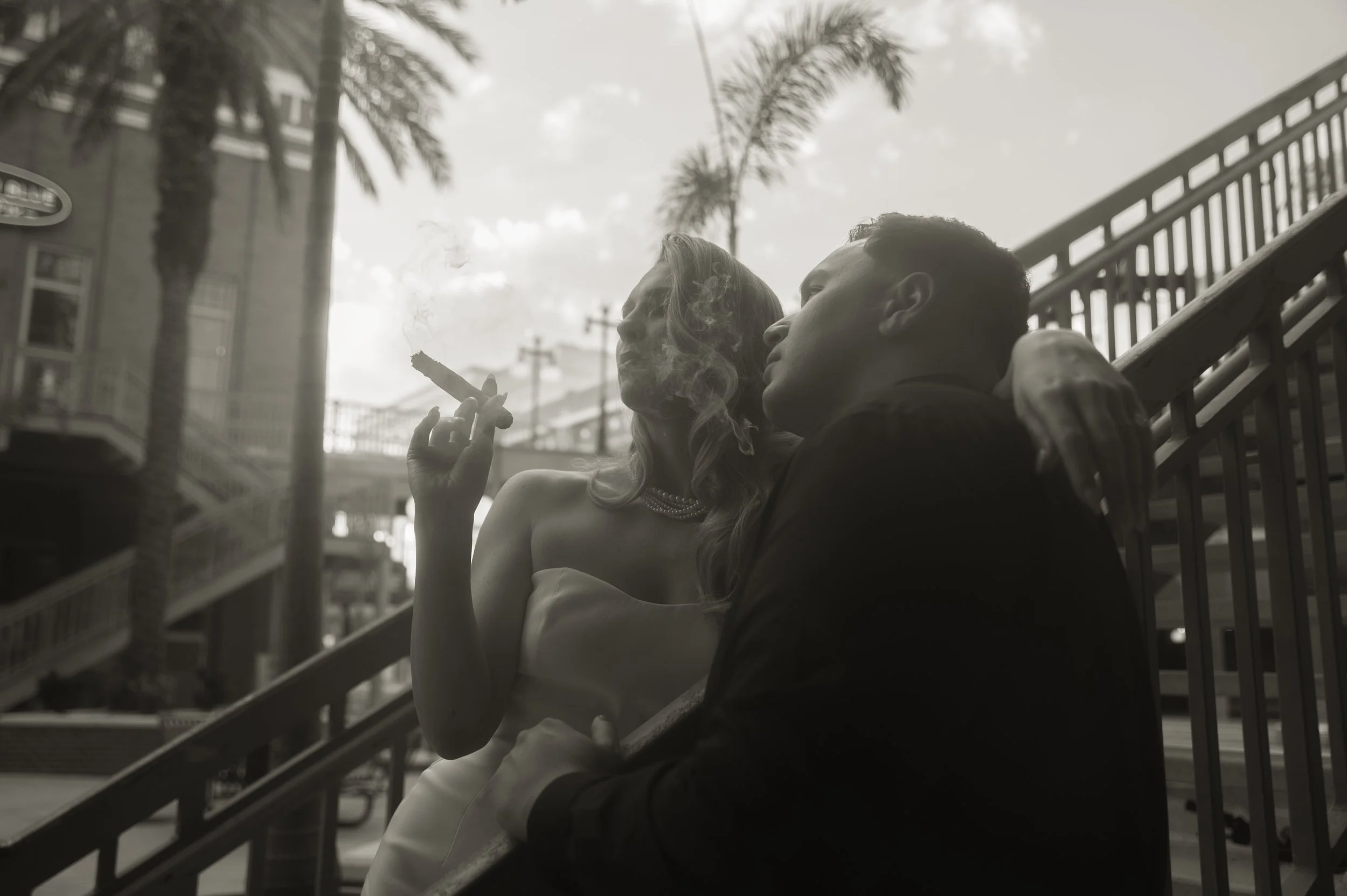 A man and woman sit close together on stairs outdoors, with palm trees and buildings in the background. The woman smokes a cigar and wears a strapless dress and pearl necklace, while the man leans into her with his arm around her shoulders.