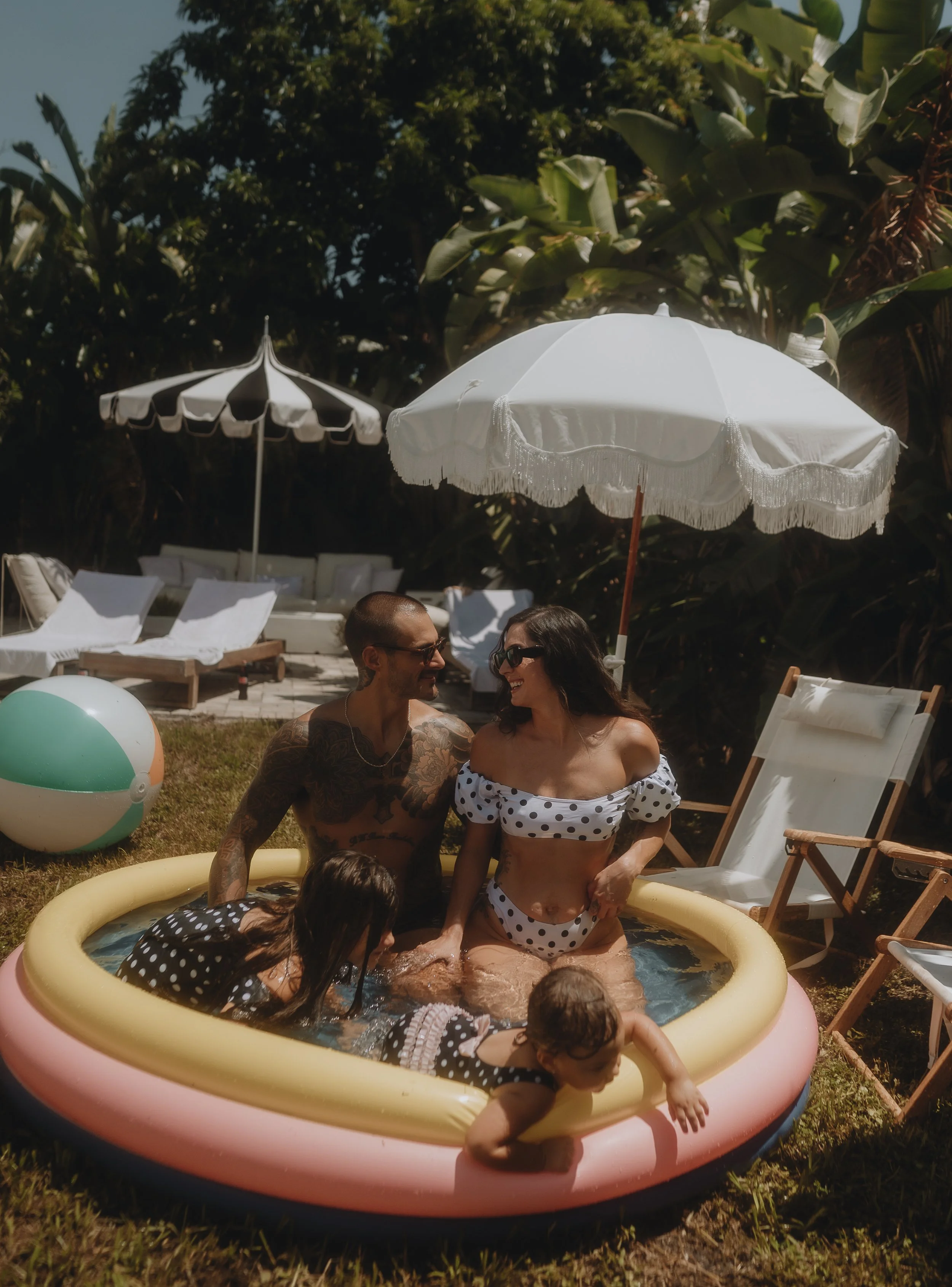 A family enjoying time together in a small inflatable pool outdoors, with a man, woman, and two children wearing swimwear. The scene is shaded by two white umbrellas, with lawn chairs and a beach ball nearby, and lush greenery in the background.
