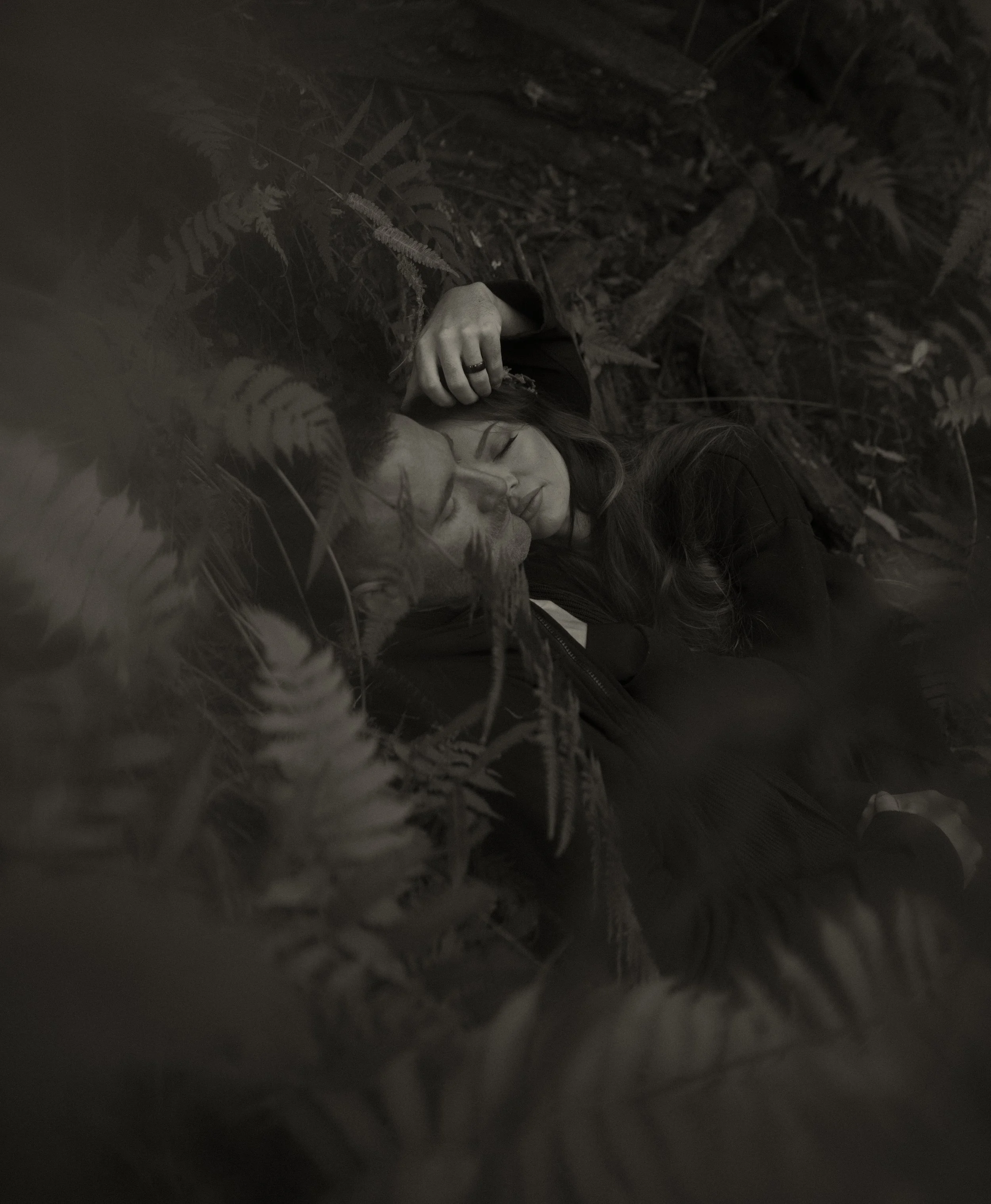 A black-and-white photo of a man and woman lying close together on the ground amongst ferns and foliage, appearing to be sleeping or resting with eyes closed.