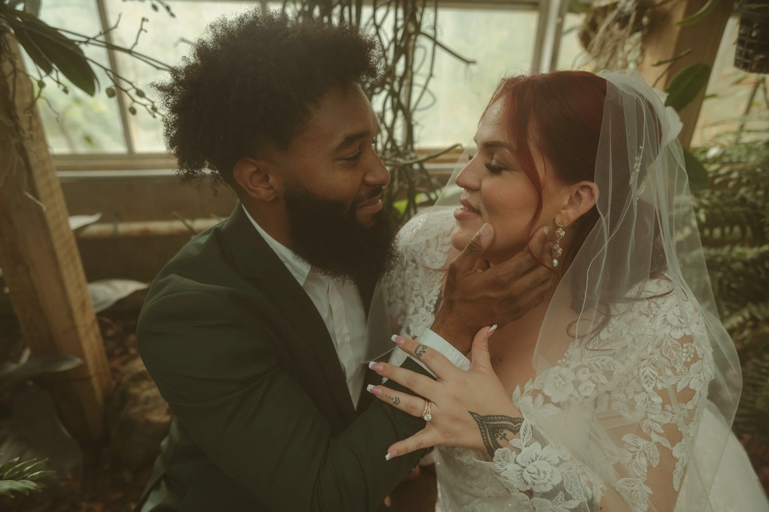 A couple on their wedding day in an intimate moment, with the man gently holding the woman's chin and looking into her eyes, while she smiles with her eyes closed, wearing a lace wedding dress and veil, in a lush greenery setting.