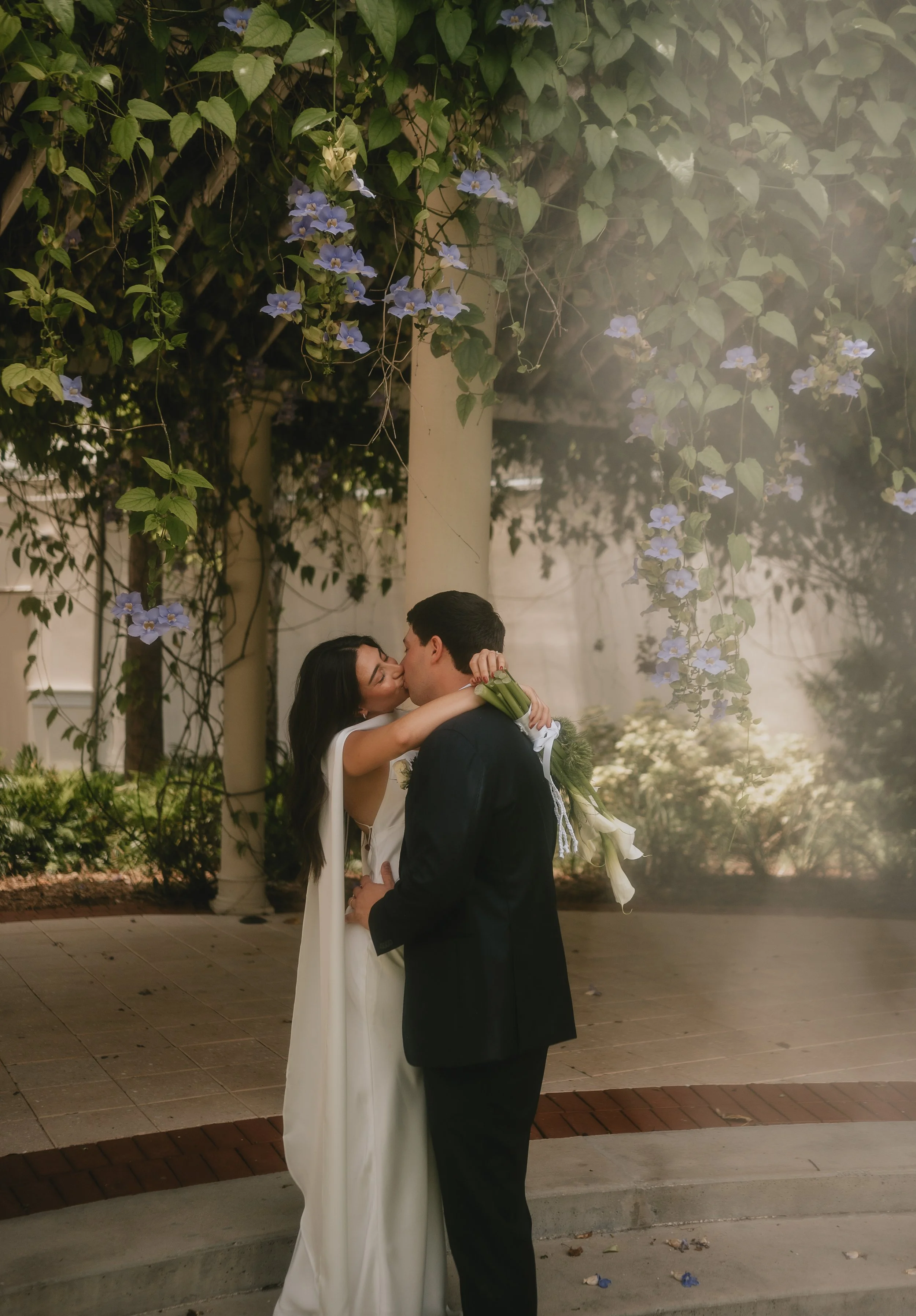 A couple kissing, the woman in a white dress with her arms around the man's neck, and the man in a dark suit holding a bouquet of white flowers, outdoors near a flowering vine-covered structure.
