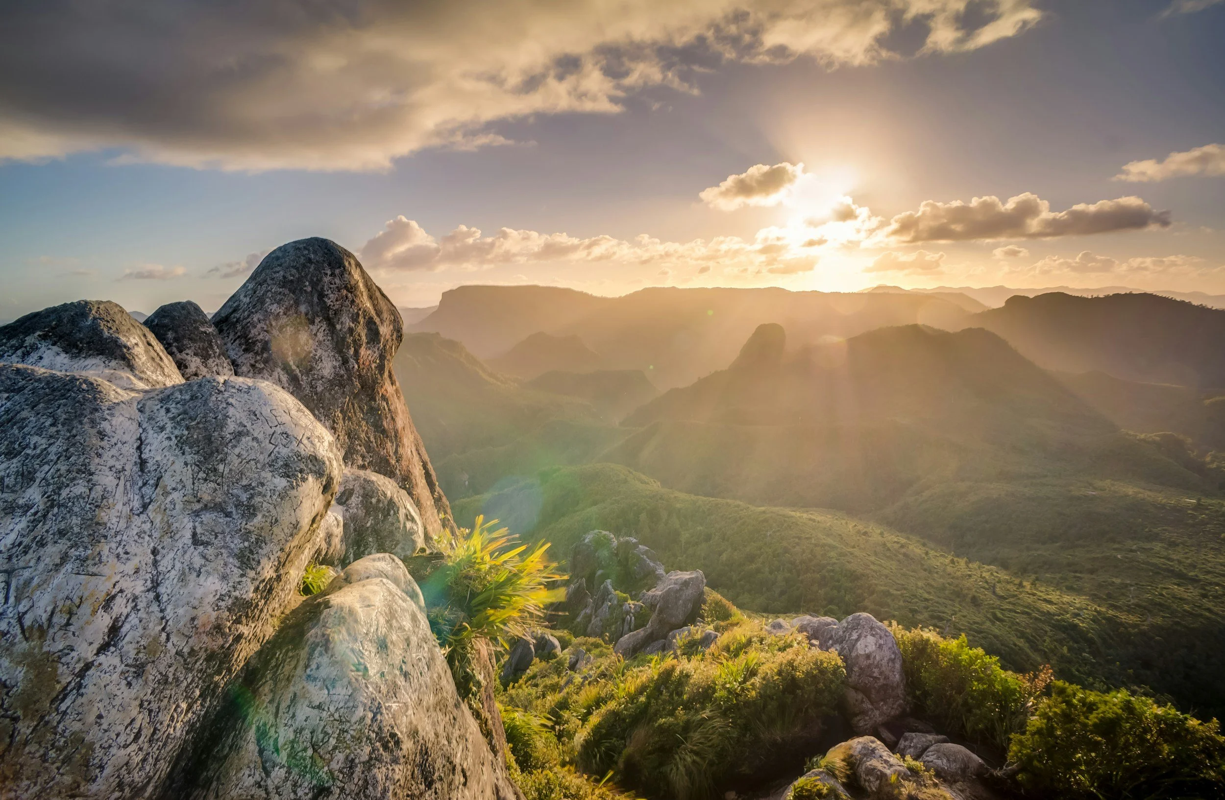 Sunset over a mountainous landscape with rocky foreground and clouds in the sky.