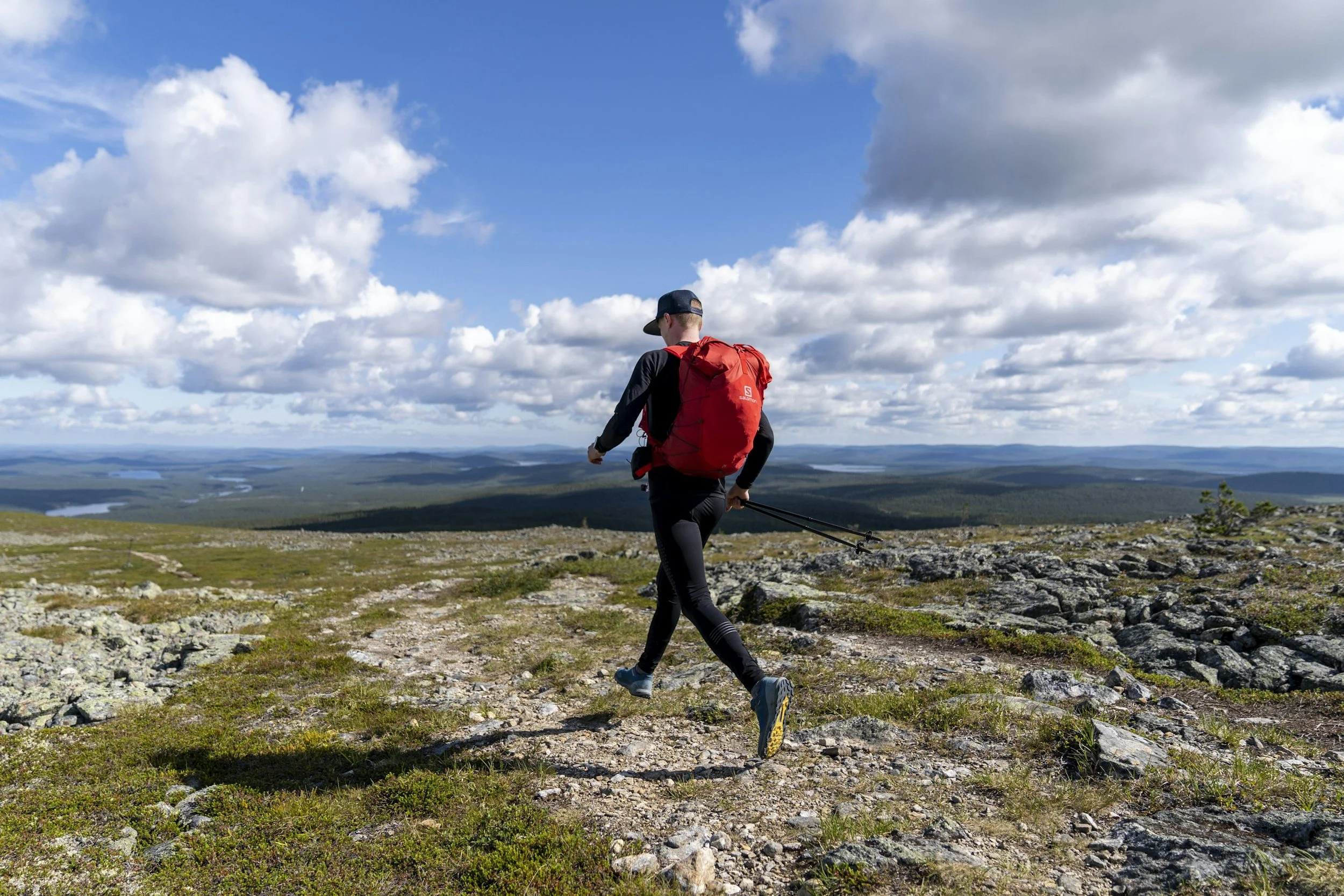 A person hiking on a rocky trail in a mountainous area with a landscape of lakes and forests in the distance under a partly cloudy sky.