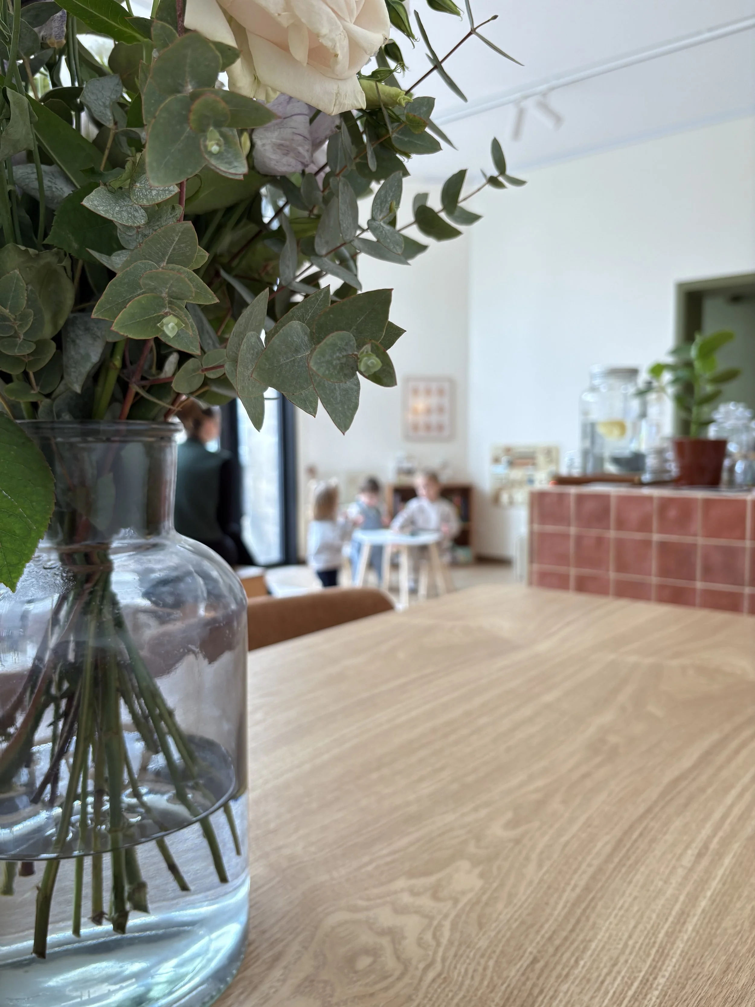Vase en verre avec des branches et des feuilles vertes, fond d'une cuisine ou d'un espace de vie avec des personnes en arrière-plan.