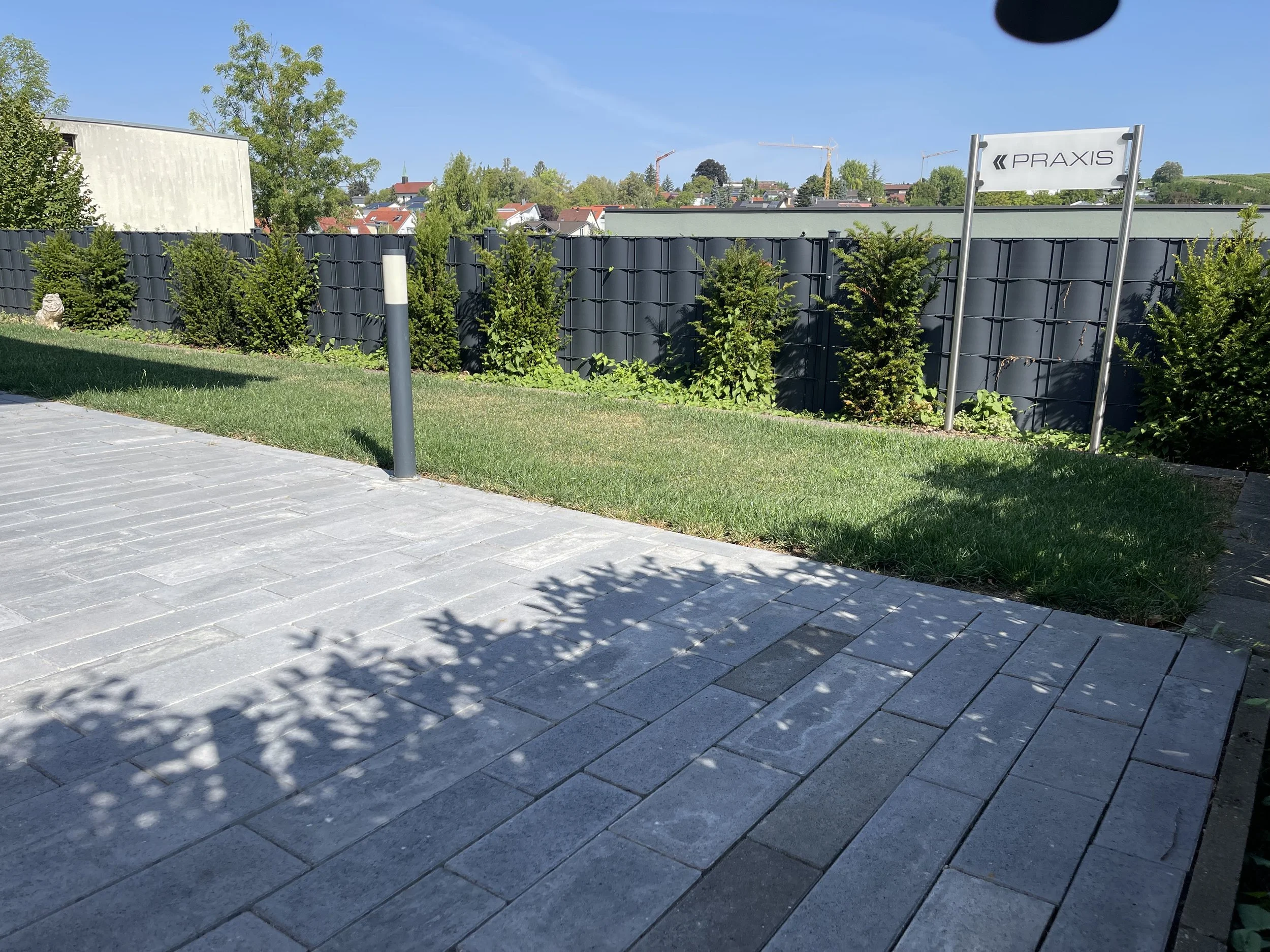 Das Bild zeigt einen gepflegten gepflegten Bereich mit einem Weg aus Fliesen, einer grünen Rasenfläche, einem dunklen Zaun, niedrigem Buschwerk und einem Orientierungsschild mit der Aufschrift 'PRAXIS'. Im Hintergrund sind Häuser, Büsche und Bäume sowie ein blauer Himmel sichtbar.