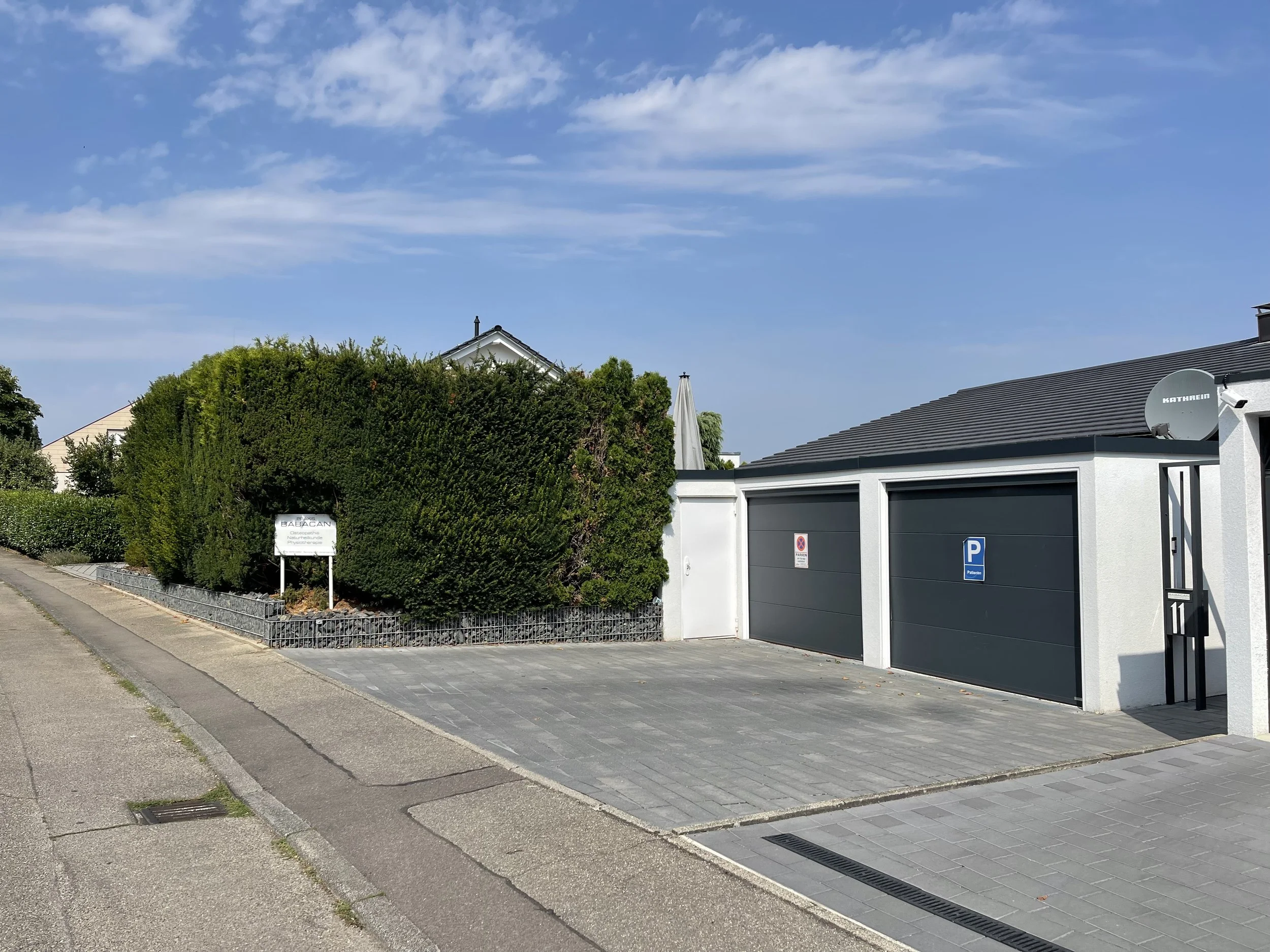 Modernes Haus mit weißen Wänden, grauem Dach, Satellitenantenne, großen Toren, umgeben von hohen Hecken, Parkpfeil und Weg in einem Vorort bei blauem Himmel.