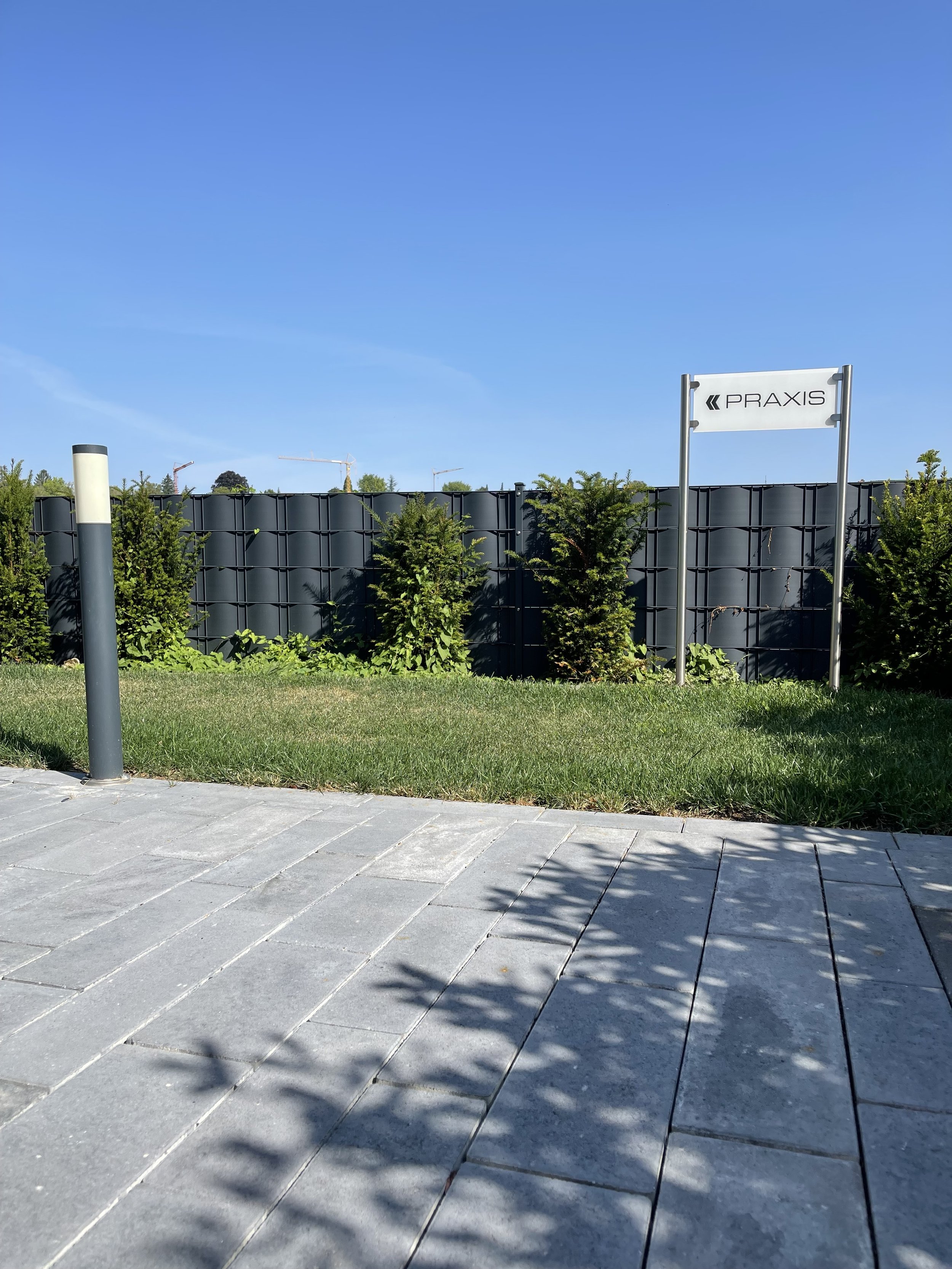 Ein heller Gehweg aus rechteckigen Platten, grüne Büsche hinter einer schwarzen Metallbegrenzung, ein blauer Himmel, ein Schild mit dem Wort »PRAXIS«. Schatten auf dem Gehweg.