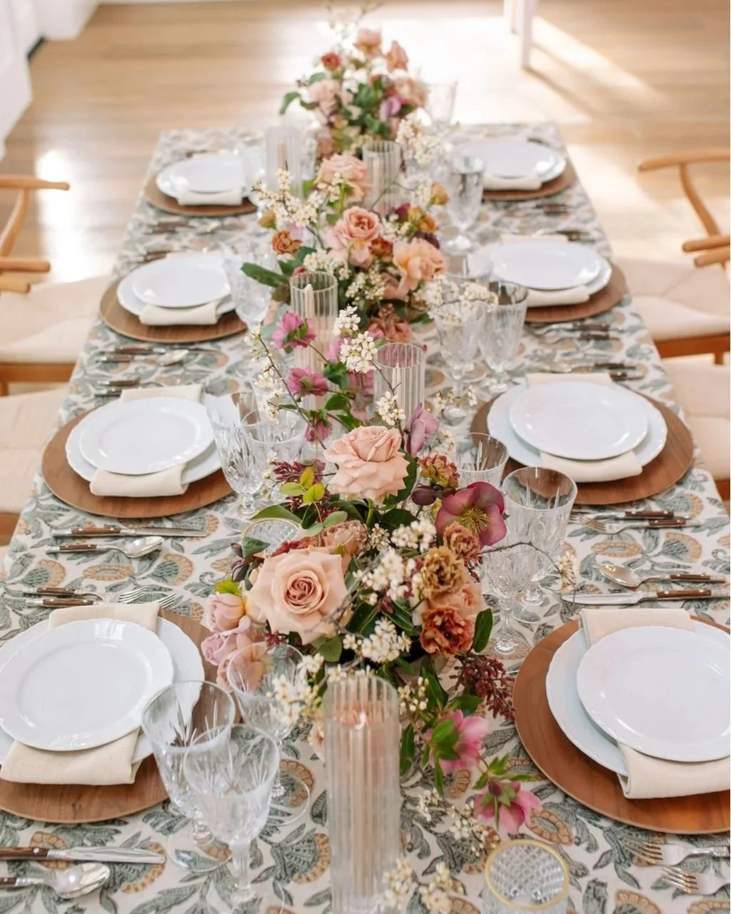 A beautifully decorated rectangular dining table with a floral centerpiece, set with white plates, crystal glasses, and silverware, surrounded by chairs, in a well-lit room.