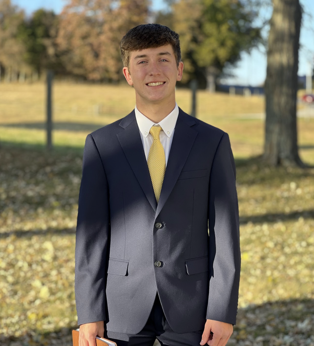 A young man in a dark suit with a white shirt and yellow tie stands outdoors on a sunny day, holding a notebook, with trees and a fence in the background.