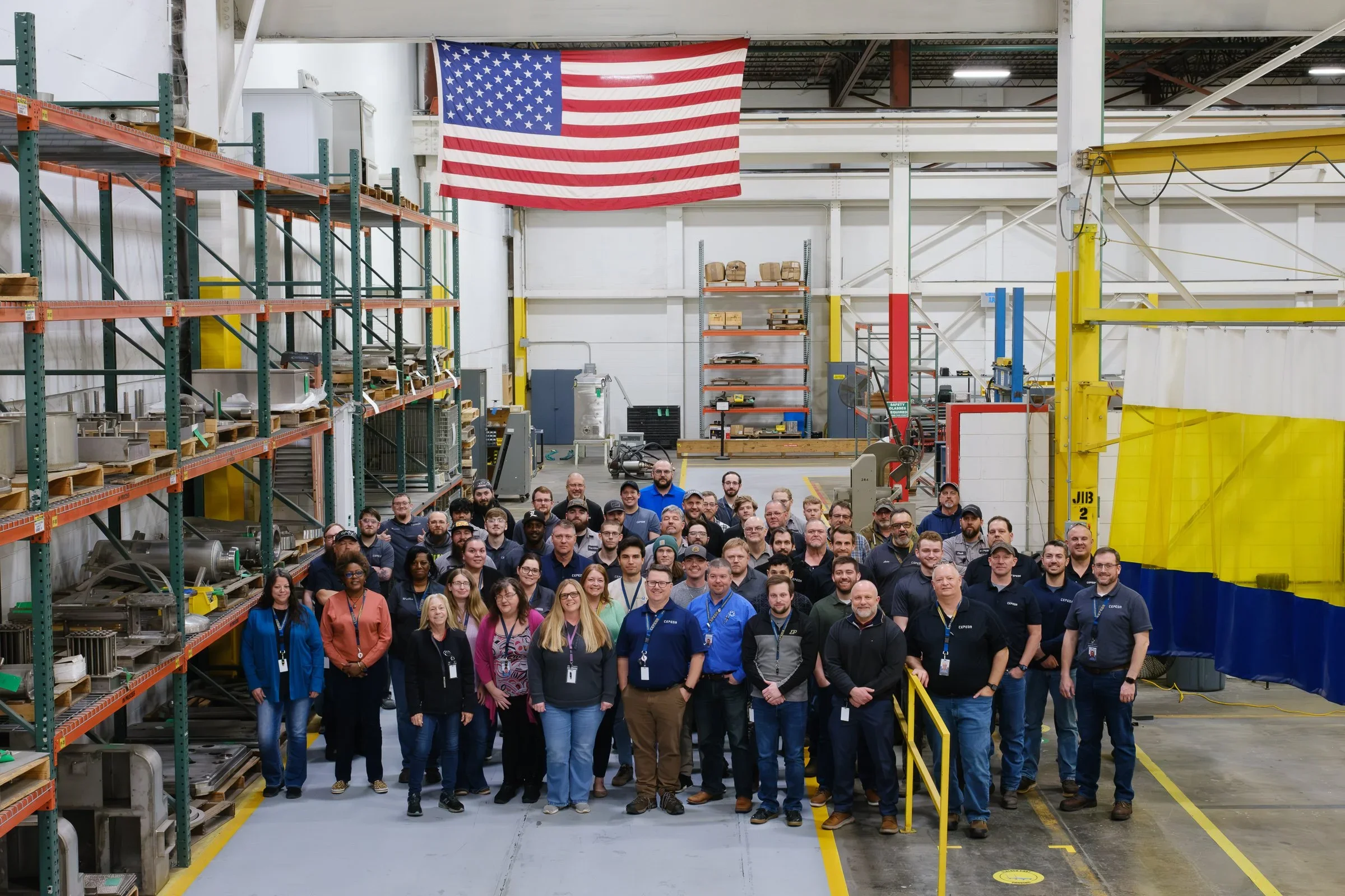 Group photo of employees standing inside a large industrial warehouse with American flag hanging above, storage racks filled with equipment and materials, yellow safety barriers, and industrial machinery.
