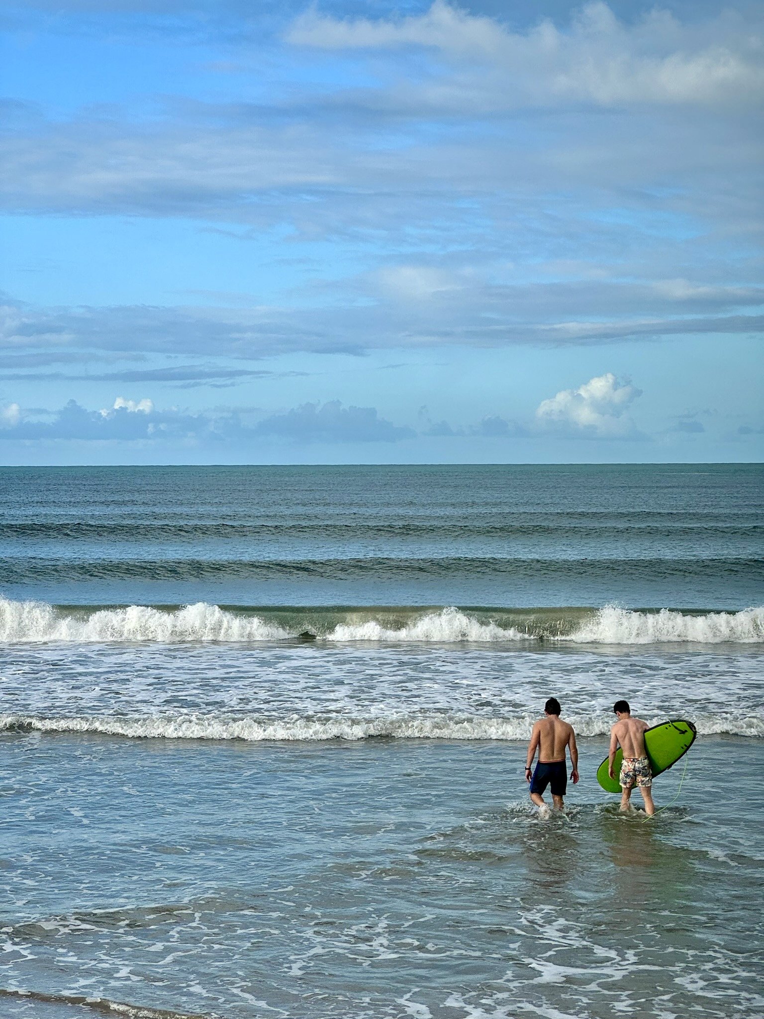 Two men walking into the ocean with a surfboard under one arm on a beach, with blue skies and clouds above