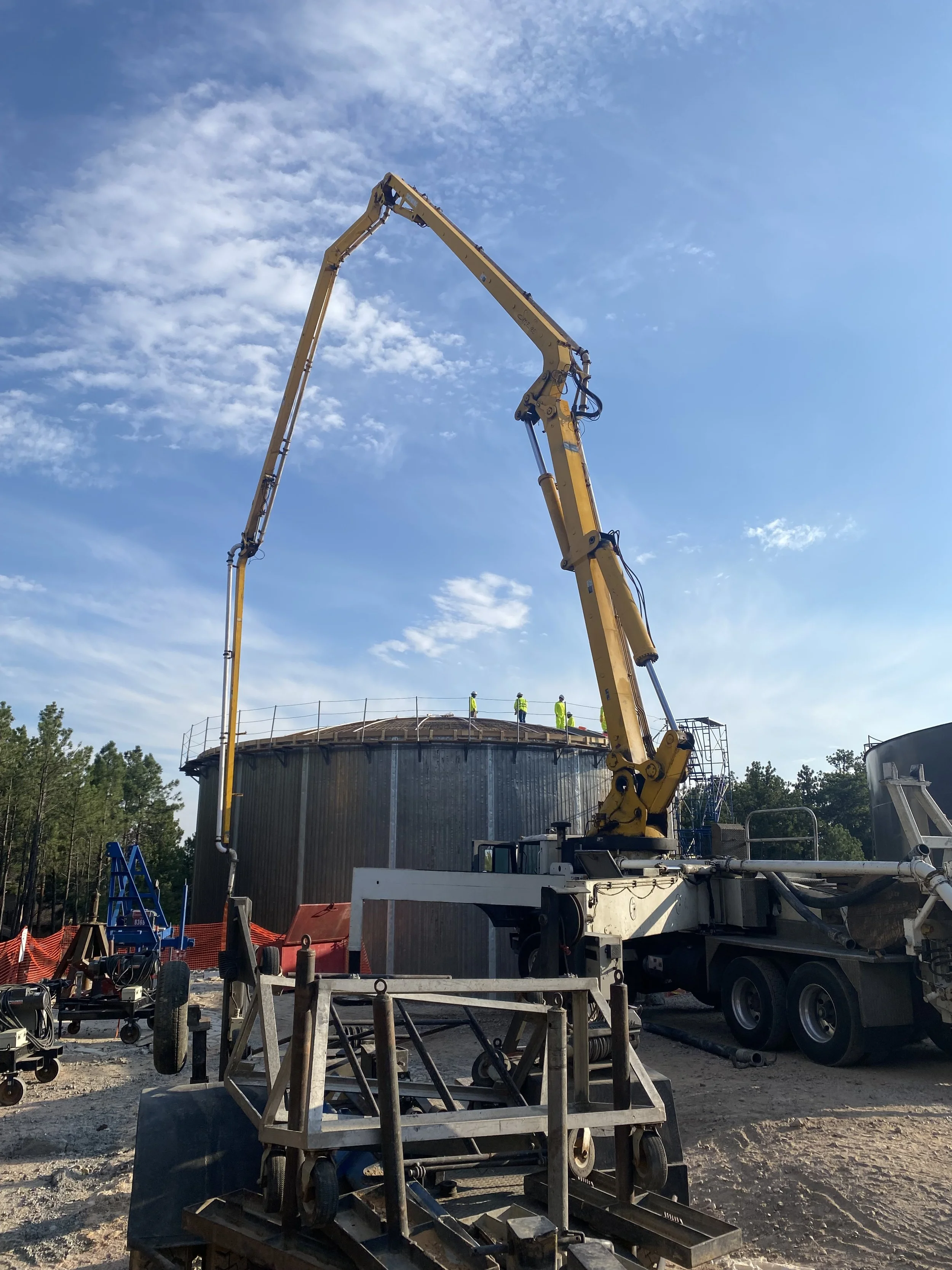 A construction site with a large concrete pump truck extending its arm over a circular metal structure, with four workers in safety gear on top, against a blue sky with scattered clouds.