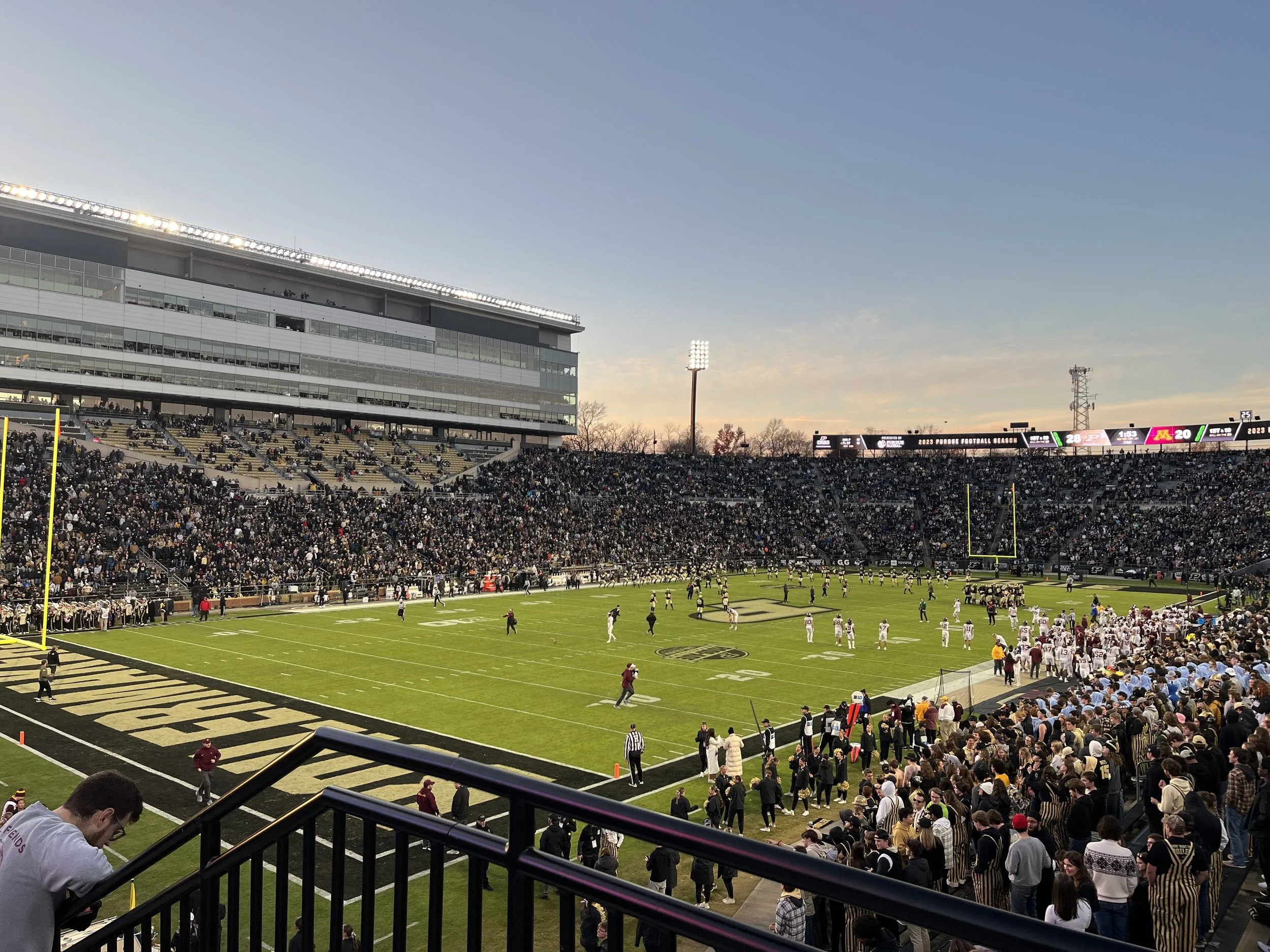 A football stadium filled with spectators during a game at dusk, with players on the field and fans in the stands.