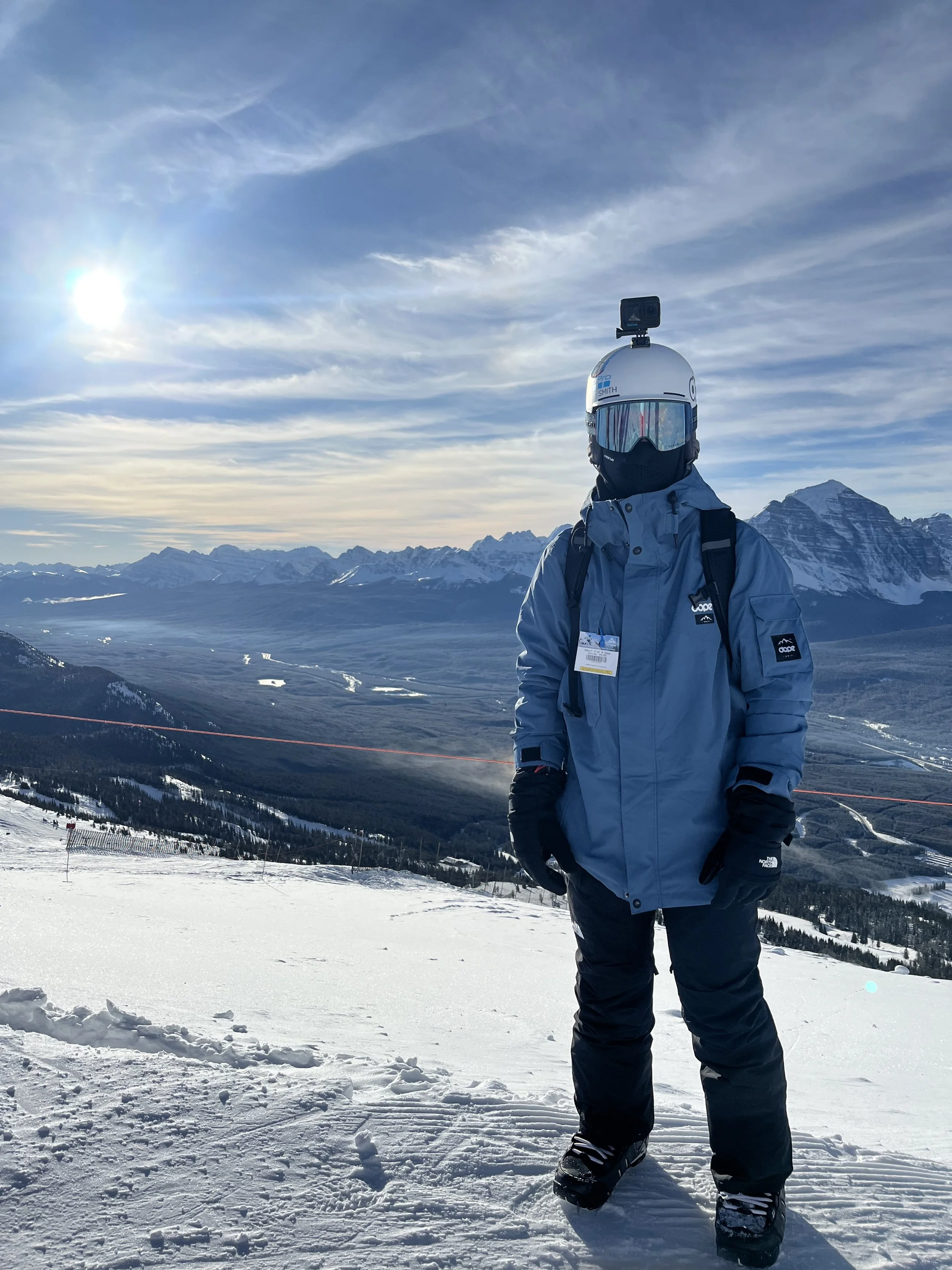 Person in ski gear standing on snowy mountain slope with mountain range and cloudy sky in the background, wearing a helmet with a camera and reflective goggles.