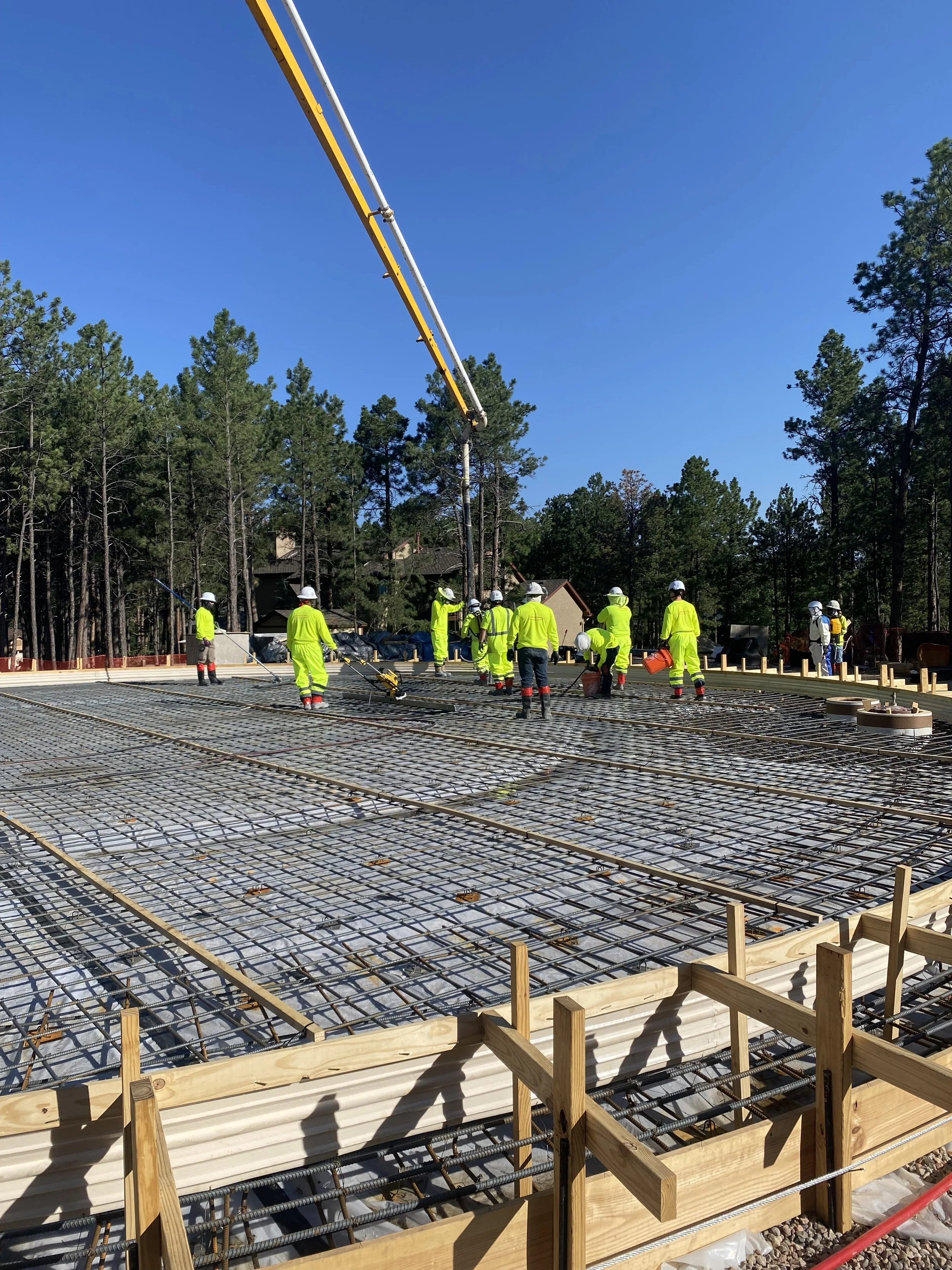 Construction workers in high-visibility clothing and hard hats working on a concrete slab with rebar reinforcement on a building site surrounded by trees.