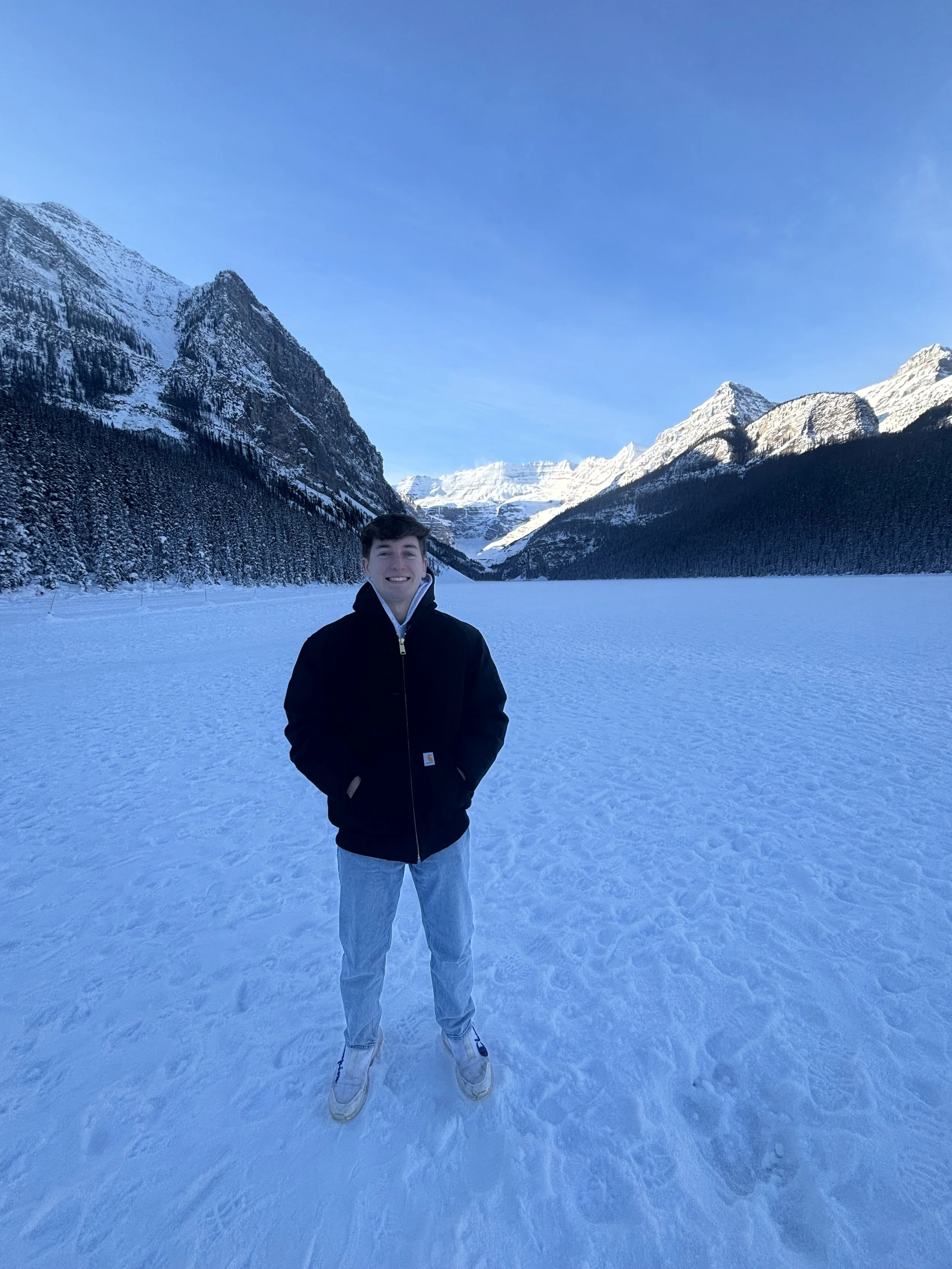 A young man standing on a snowy landscape with snow-covered mountains and a blue sky in the background.