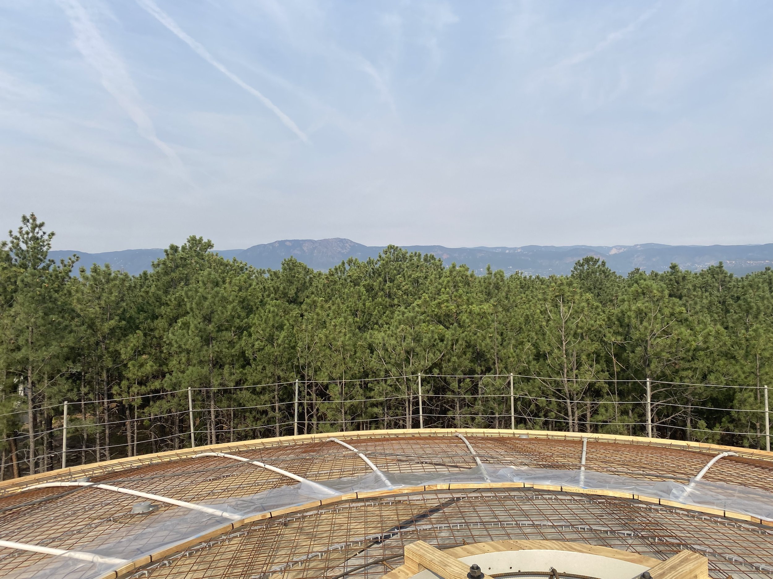 View from a construction site with curved rebar and wooden formwork, overlooking a green forest with mountains in the distance under a blue sky.
