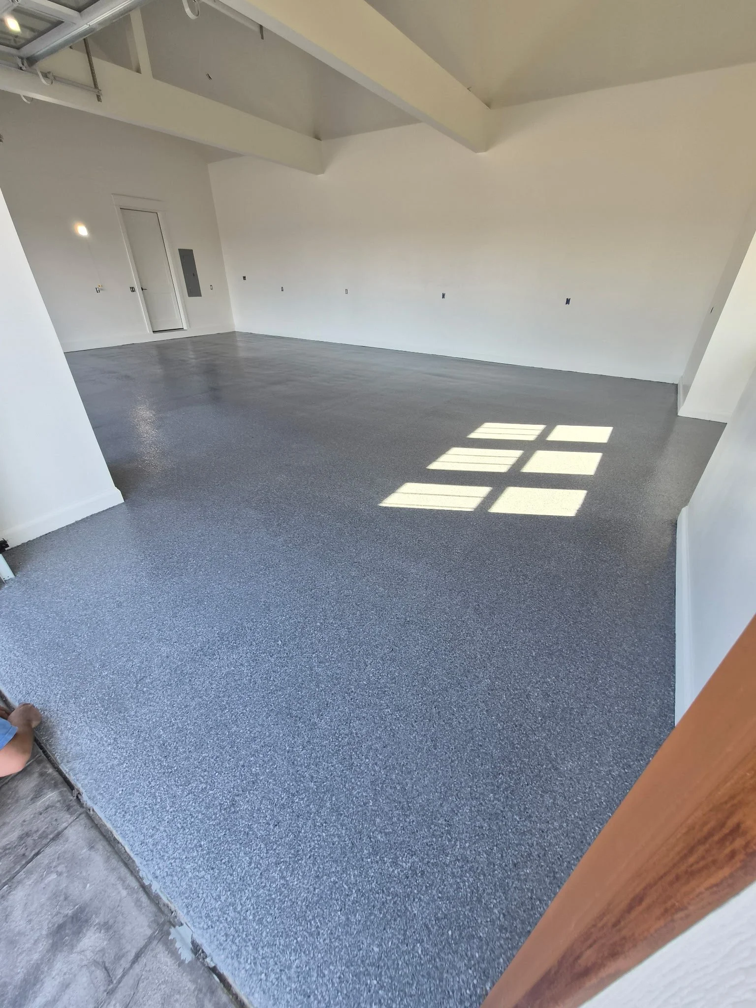 Empty room with gray epoxy flooring, white walls, and a window allowing sunlight to cast a grid pattern on the floor.
