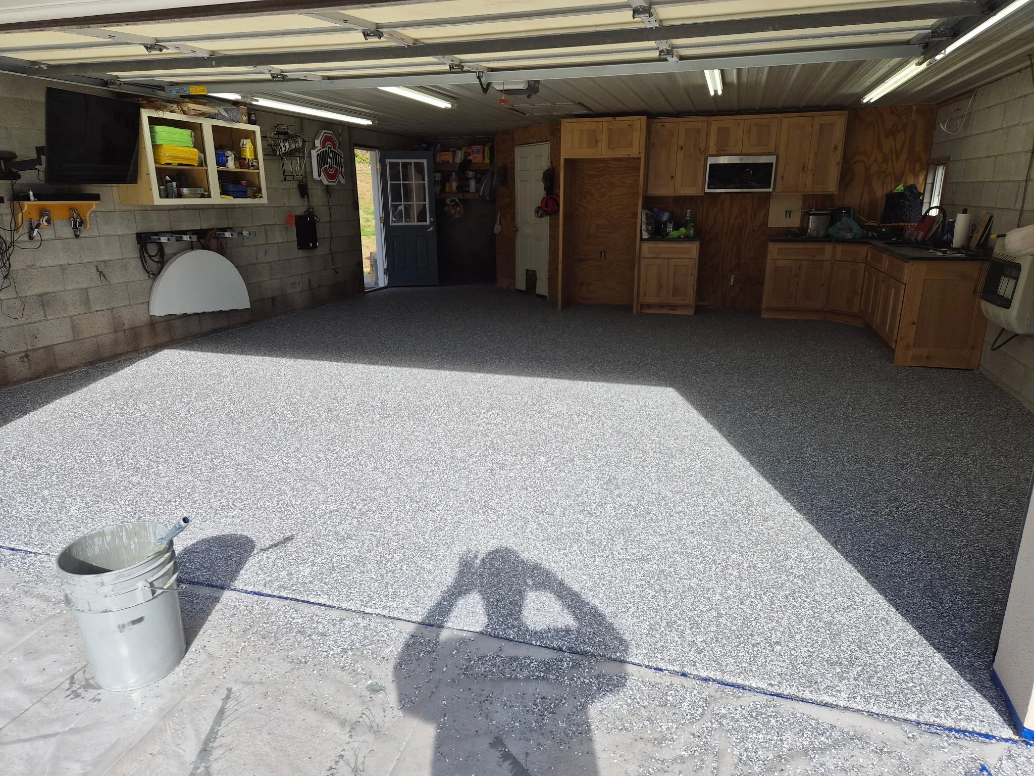 Empty garage interior with a newly coated speckled epoxy floor, wooden cabinets on the right and shelves on the left, open door at the back, and the shadow of the person taking the photo