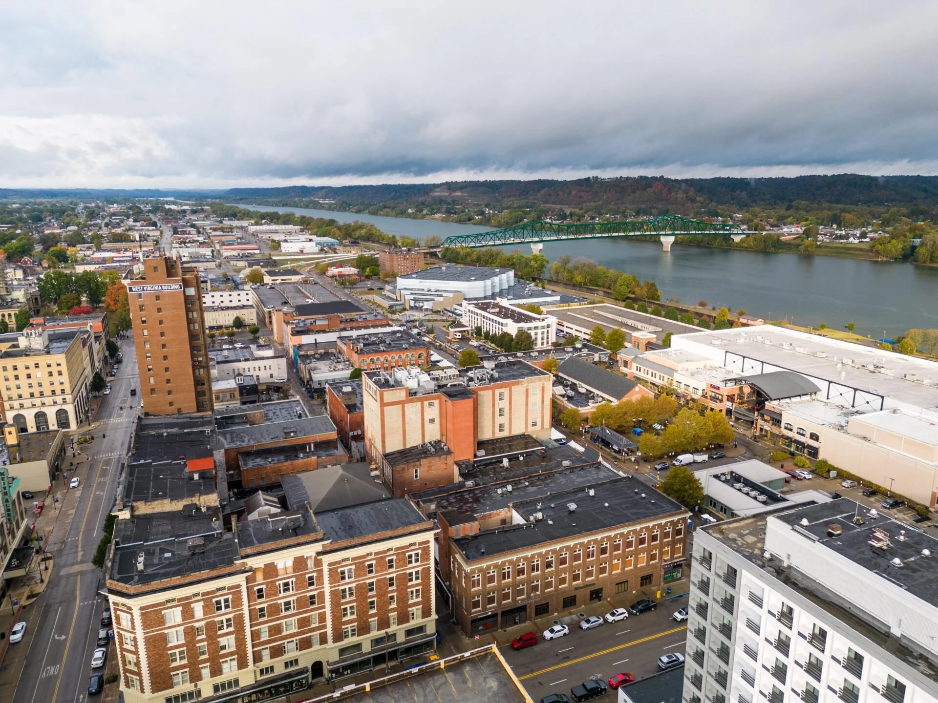 Aerial view of Huntington, WV with the Ohio River in the background, featuring various buildings, streets, and a bridge crossing the river under cloudy skies.