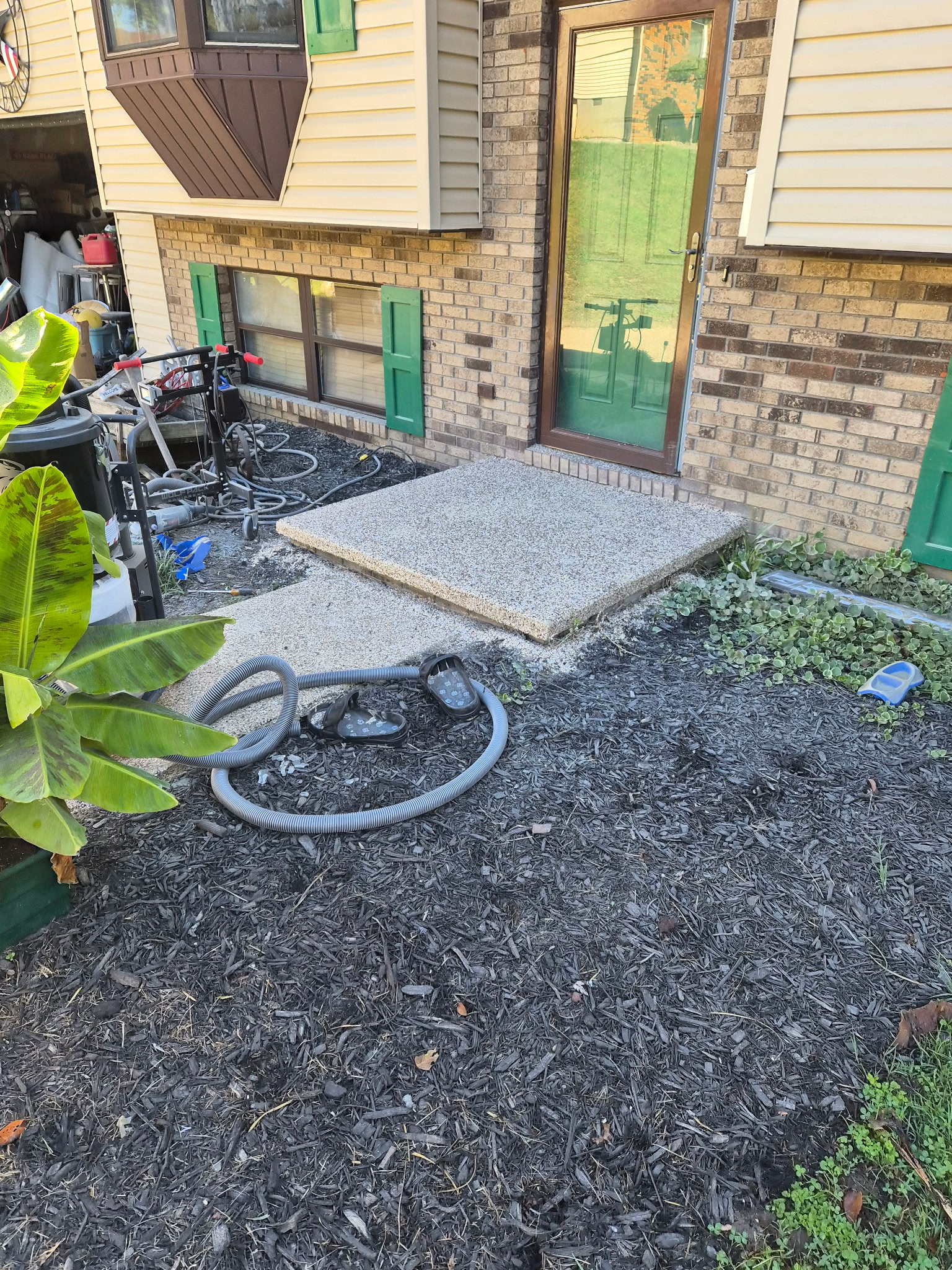 Backyard patio area with a concrete step, a glass door, and a window with green shutters. There are gardening tools and hoses, and some newly planted small plants along the border.