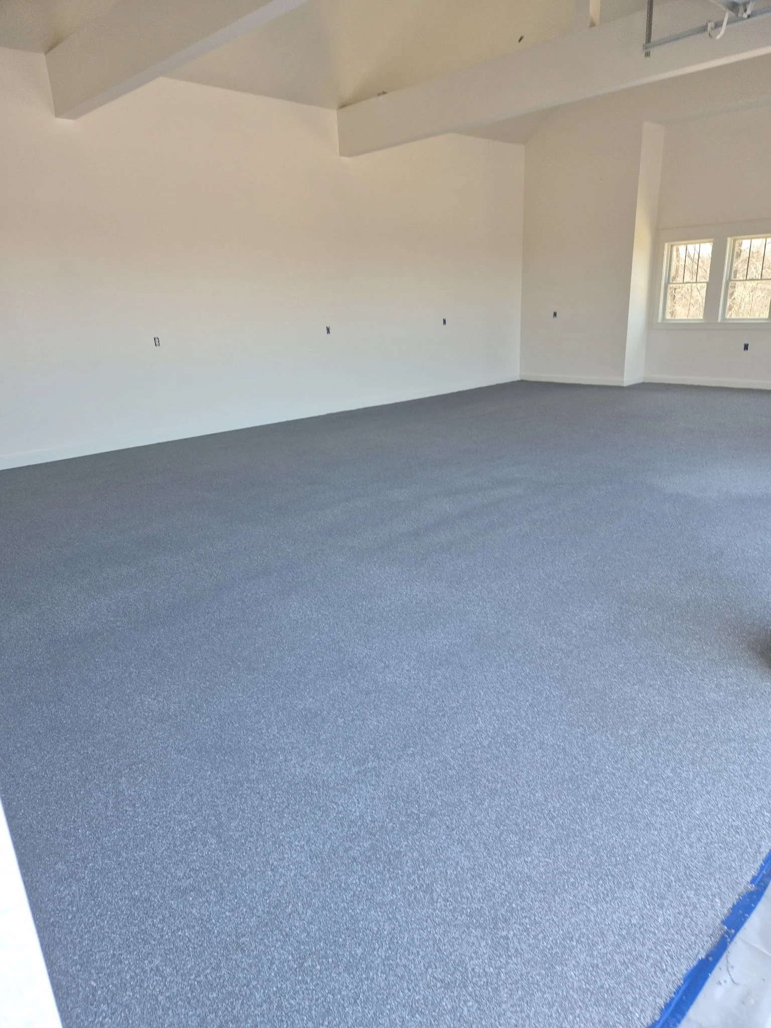 Empty room with white walls, several electrical outlets, and a large window with blinds. The room has a dark speckled carpet and visible ceiling beams.
