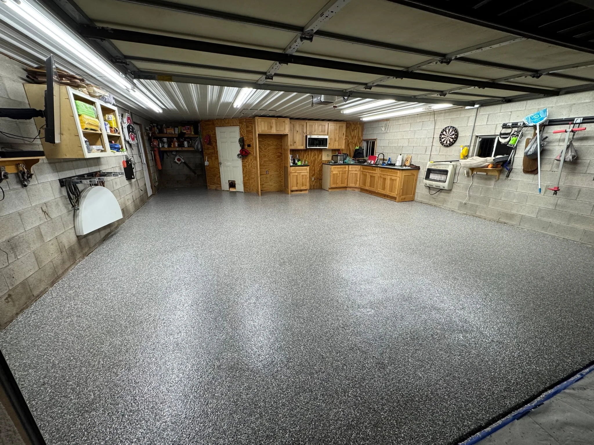 Empty garage with speckled gray epoxy floor, wood cabinets, pegboard wall with tools, and a dartboard on the back wall.