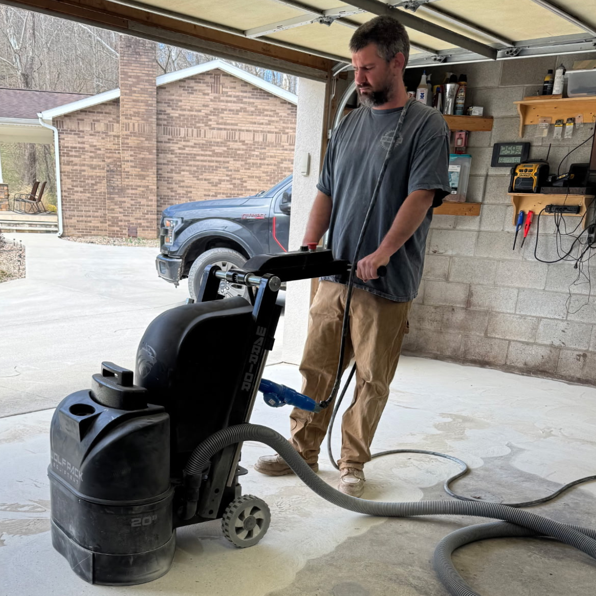 A man using a large wet/dry vacuum cleaner inside a garage, with work shelves on the wall and a driveway with a pickup truck outside.