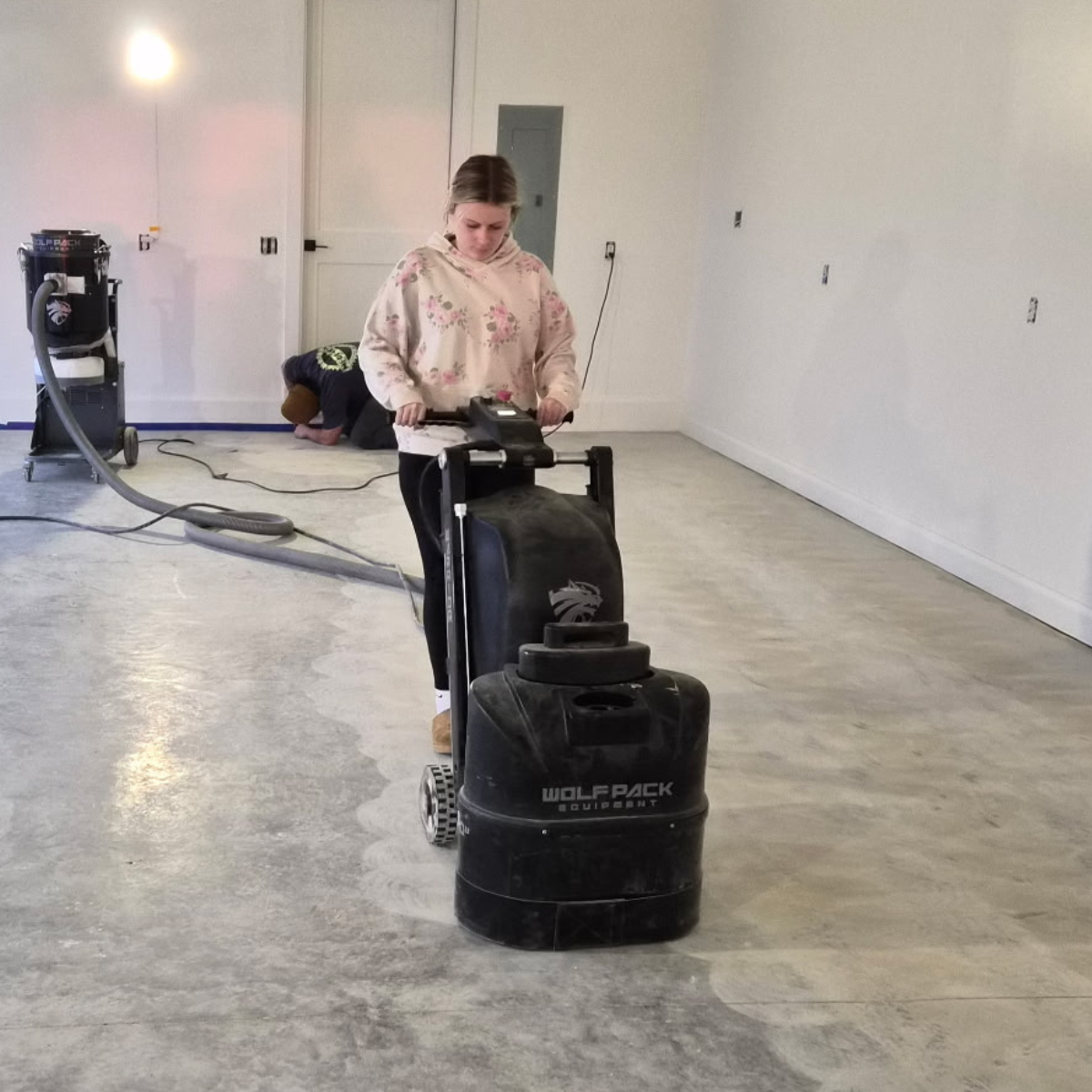 A woman operating a floor polishing machine in an empty room with white walls and a concrete floor. A person is seen in the background kneeling on the floor.